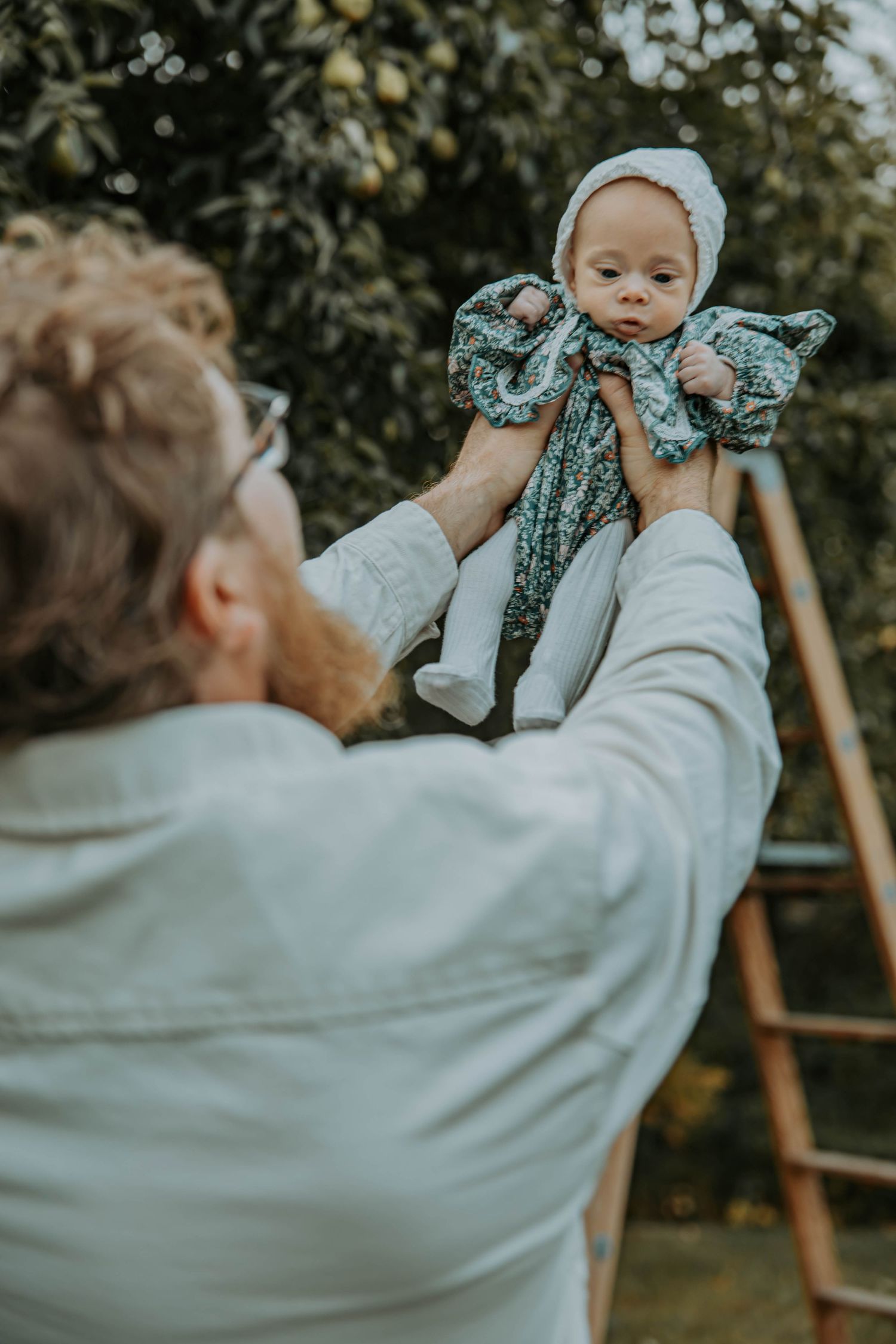 A parent lifting and playfully interacting with a baby wearing a floral bow headband against a garden background.