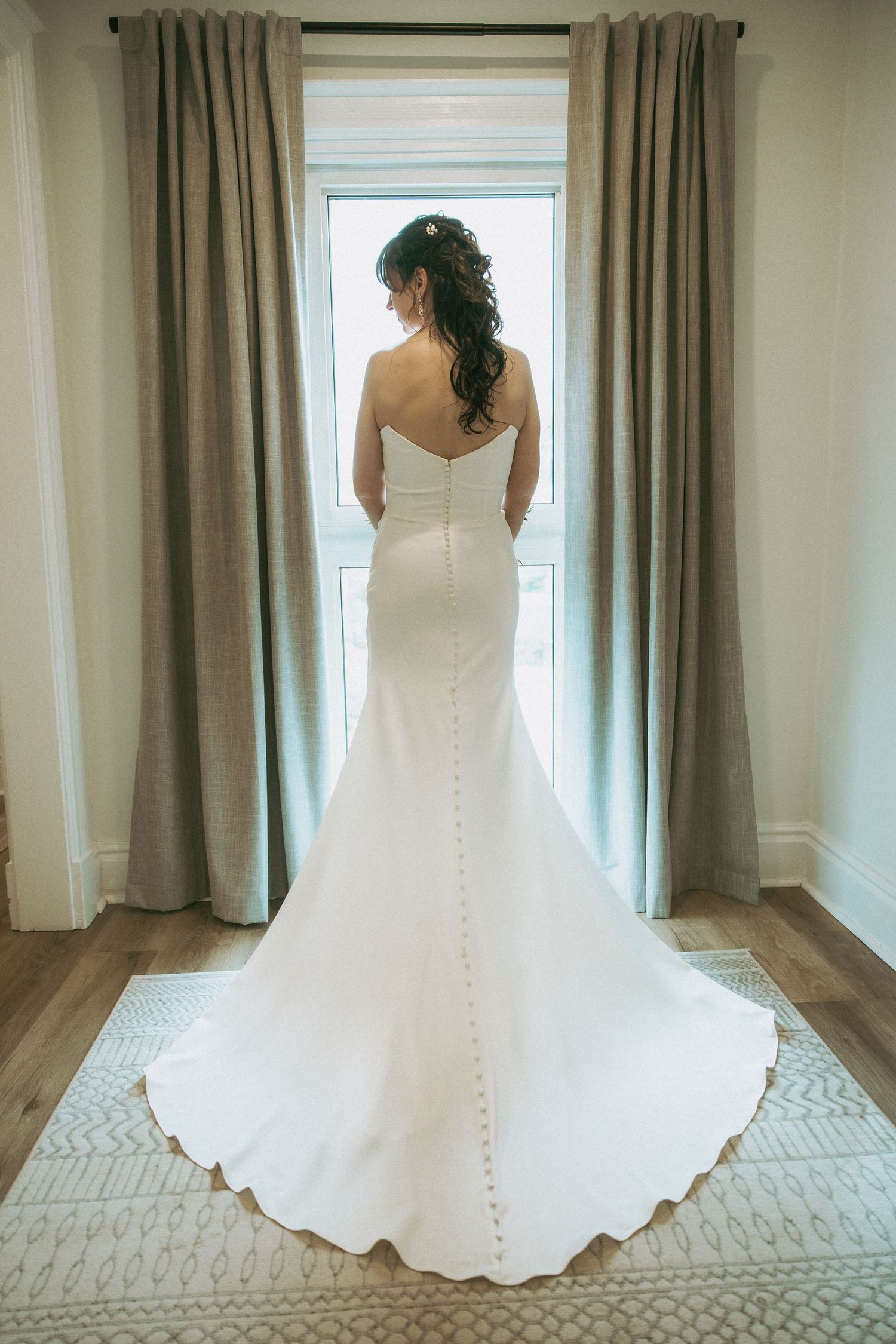 Bride in strapless white mermaid wedding gown with button train stands before a bright window with gray curtains.