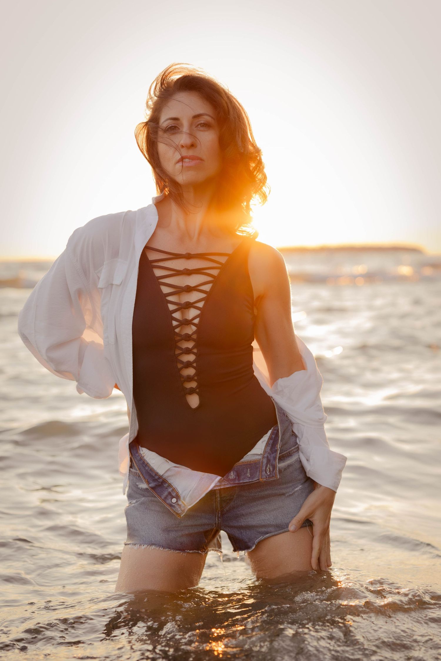 Figure in black swimsuit and white shirt standing in ocean water at sunset with warm golden backlighting.