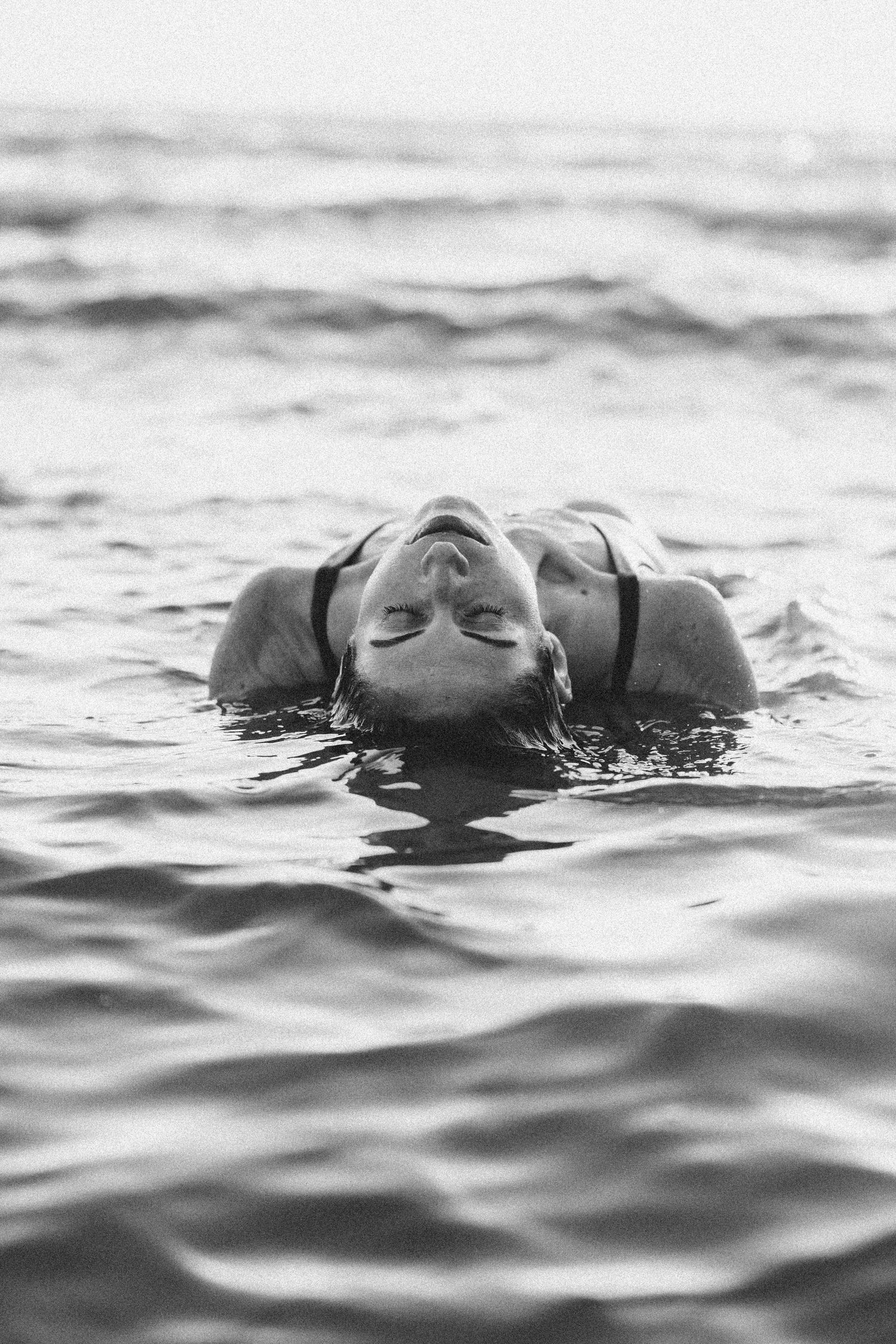 A black and white photo showing someone floating peacefully in dark rippling water with their eyes closed.