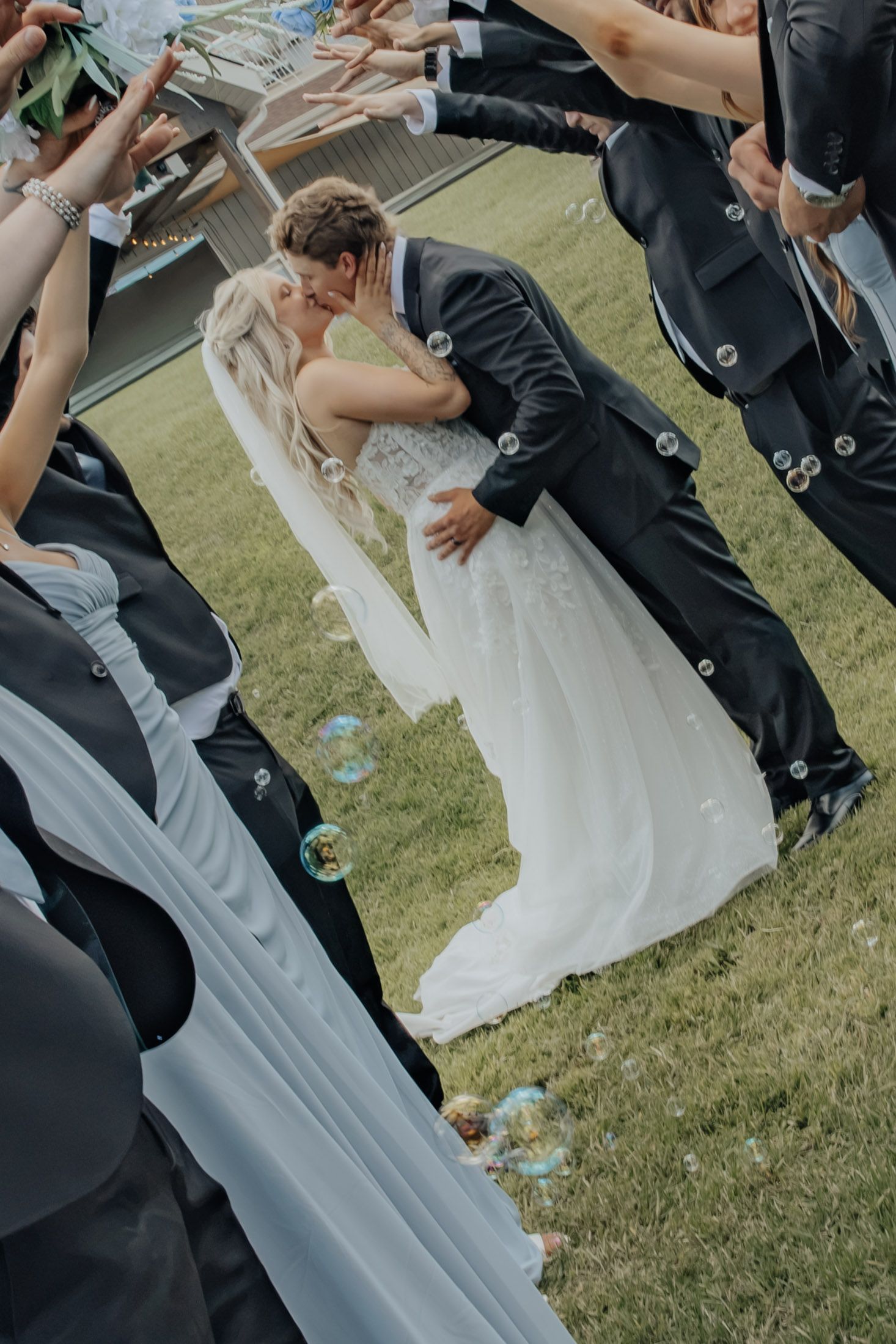 Bride and groom share a kiss surrounded by wedding party blowing bubbles on a green lawn.