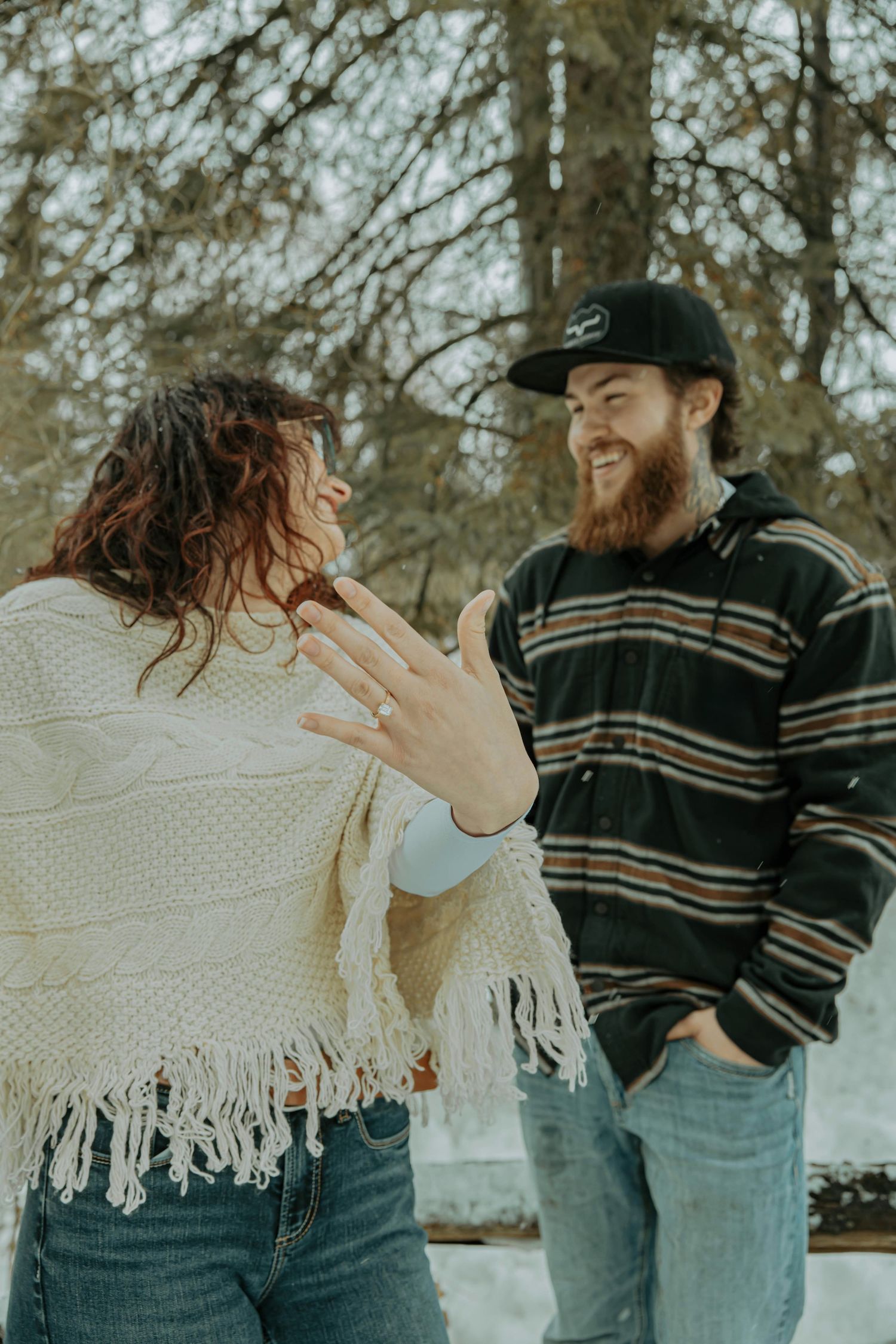 A couple shares a playful moment in the snow while wearing winter clothing and a white knit blanket.