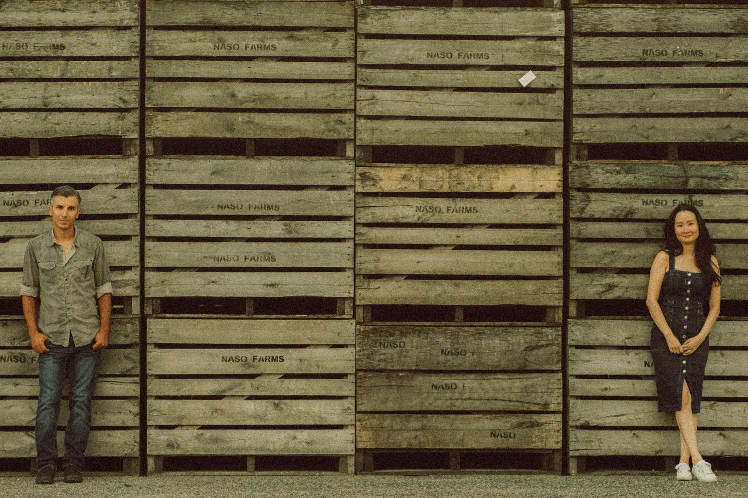 A man and woman stand apart against a rustic wooden crate wall outdoors in a casual portrait.