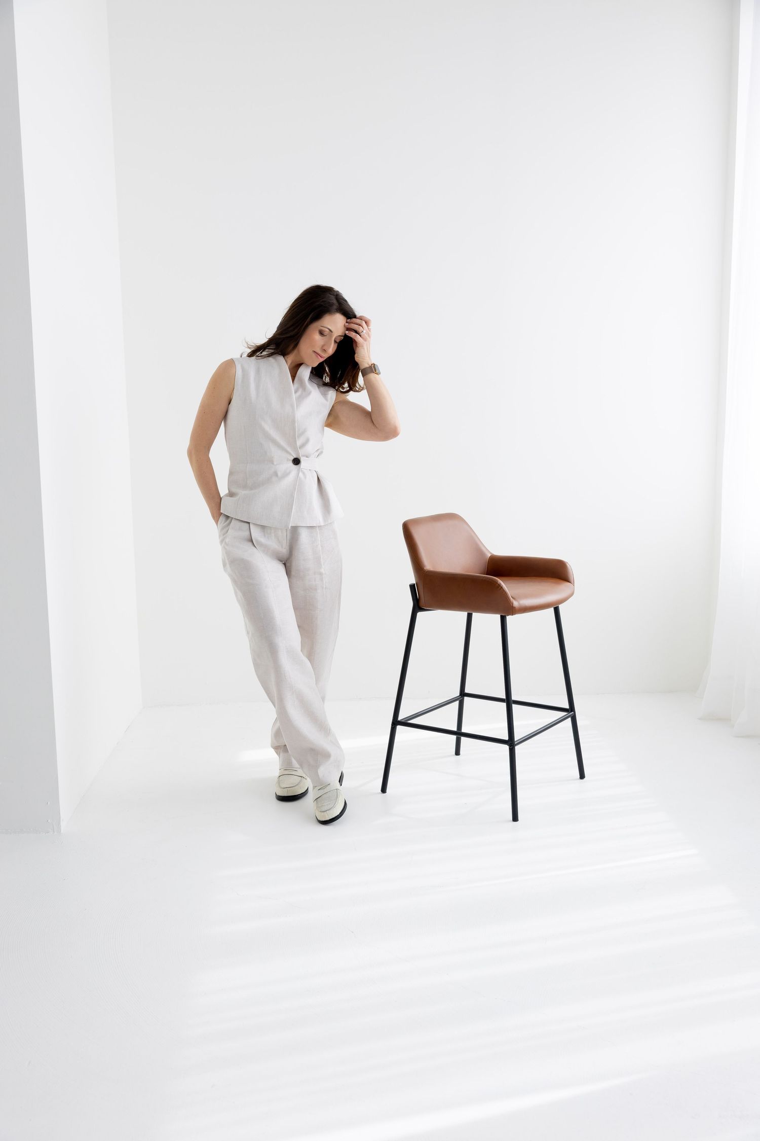 Woman in white sleeveless vest suit standing beside brown leather bar stool in minimalist white studio setting.