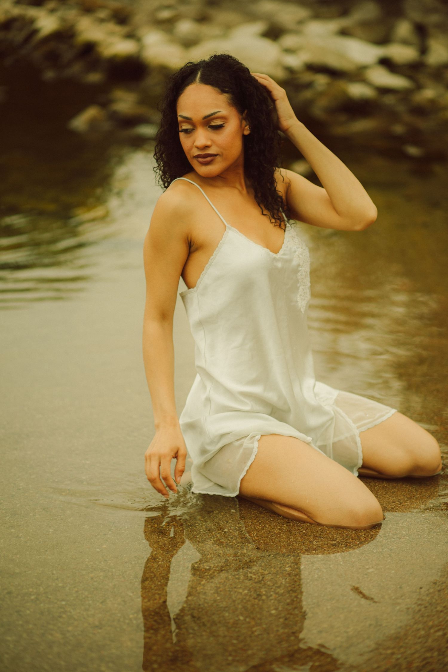 Young woman in white slip dress kneeling gracefully in shallow water near rocky shore.