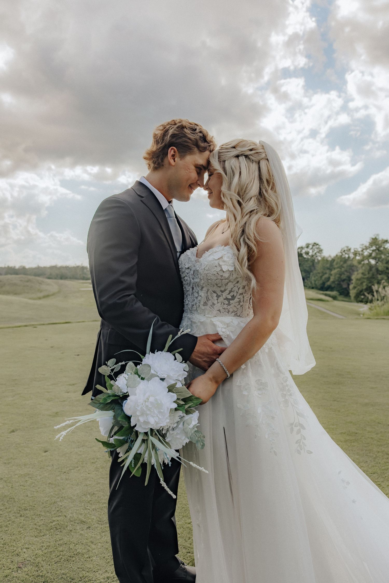 Bride and groom touching foreheads outdoors on golf course, bride holding white peony bouquet, cloudy sky backdrop.