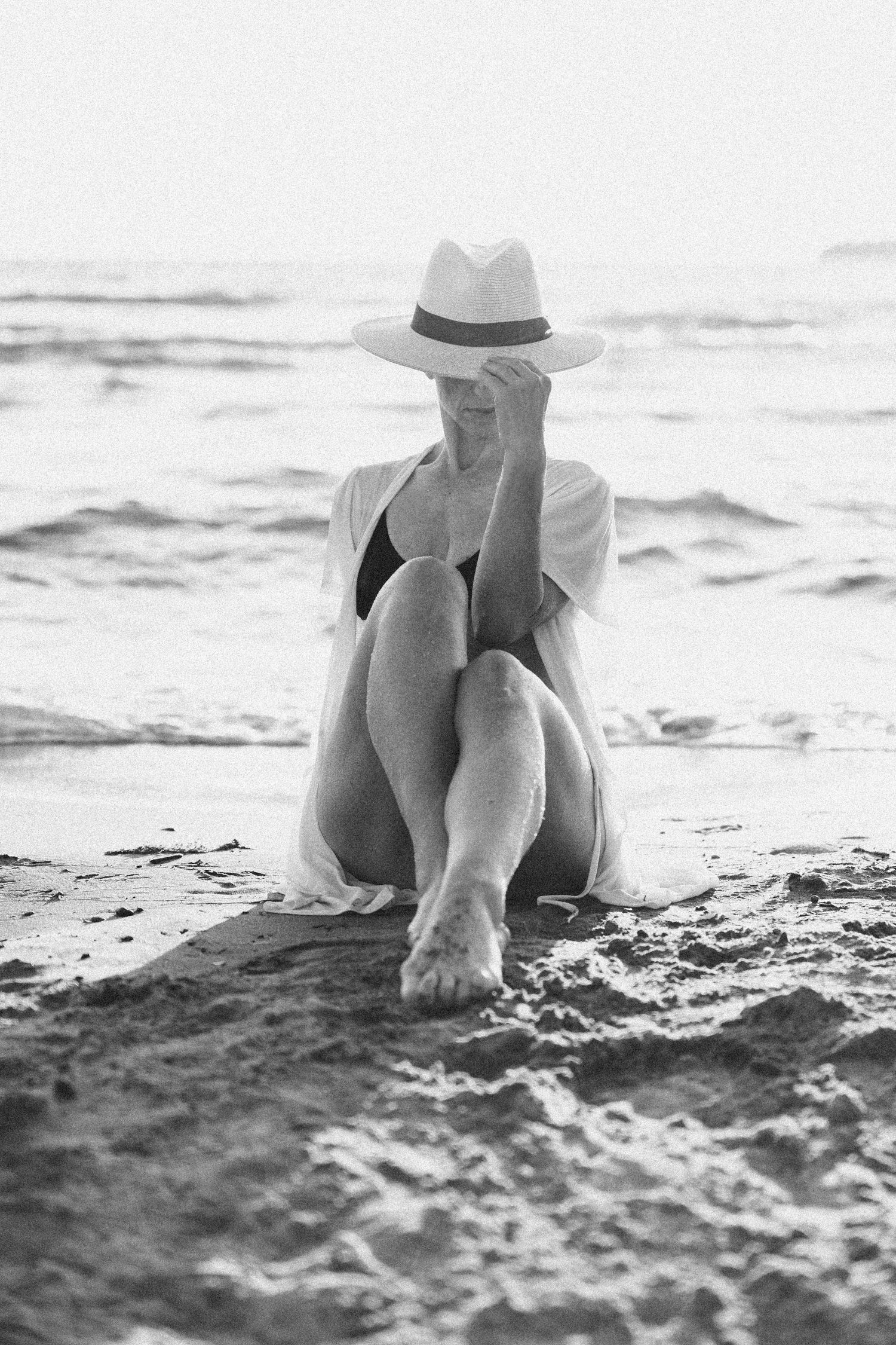 A figure wearing a white fedora hat sits alone on a sandy beach in a black and white artistic photograph.