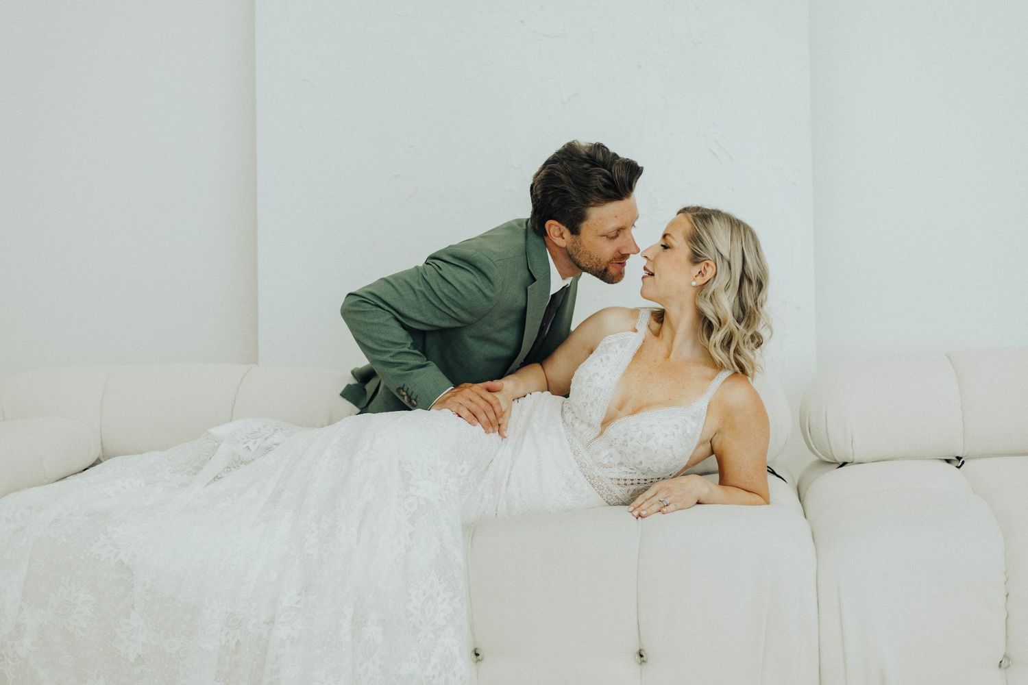 A couple in wedding attire share an intimate moment while lounging on a white couch together.