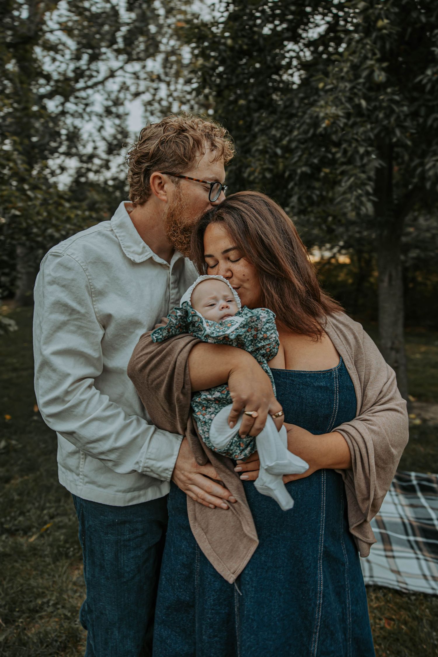 Family shares tender outdoor moments together while sitting on a plaid blanket in a wooded setting.