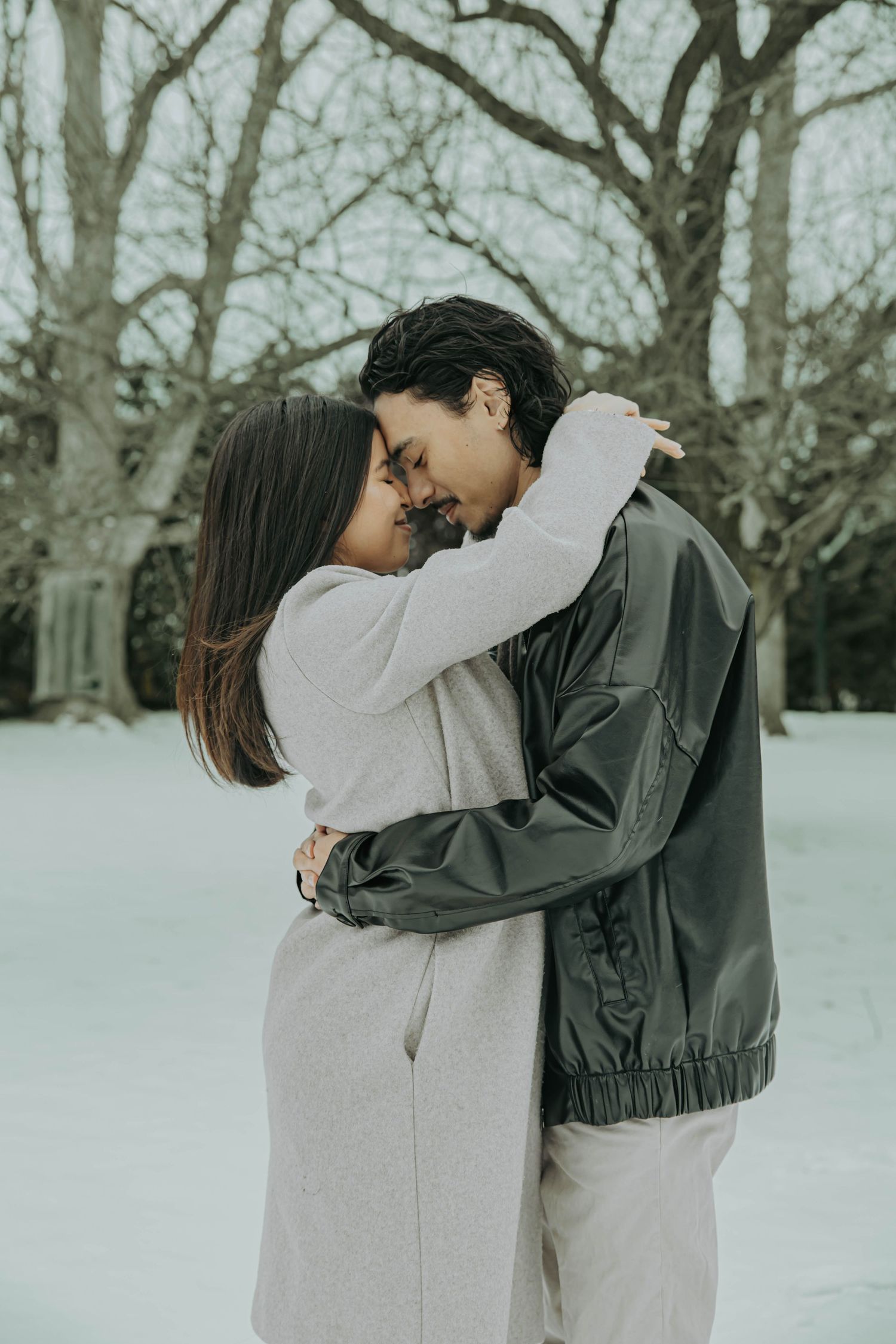 A couple embraces in a tender moment against a snowy winter backdrop with bare trees in the background.