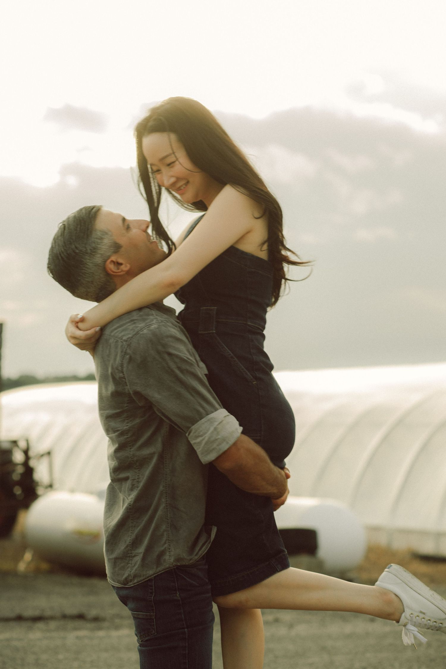 A man lifts a smiling pregnant woman outdoors, sharing a joyful romantic moment against a cloudy sky.