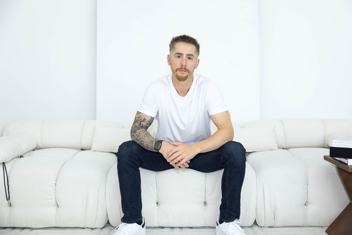 Person in white t-shirt and dark pants sitting casually on white couch in bright minimalist room.