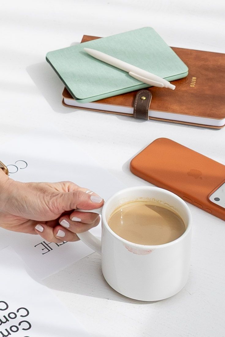 A hand holding a white coffee mug beside leather notebooks, a mint journal, pen, and orange phone case on a white surface.