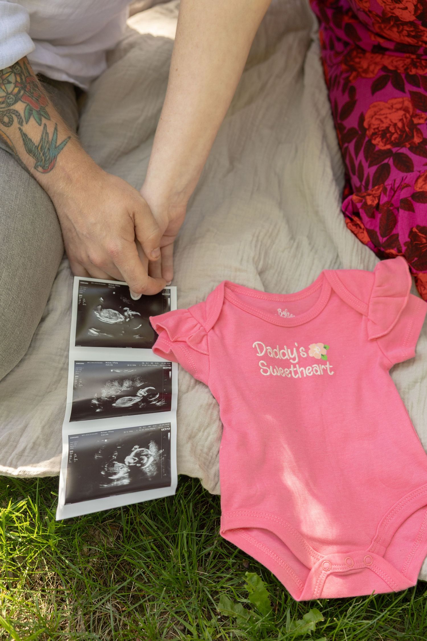 Ultrasound photos and pink Daddy's Sweetheart baby onesie on blanket, held by tattooed hands outdoors.