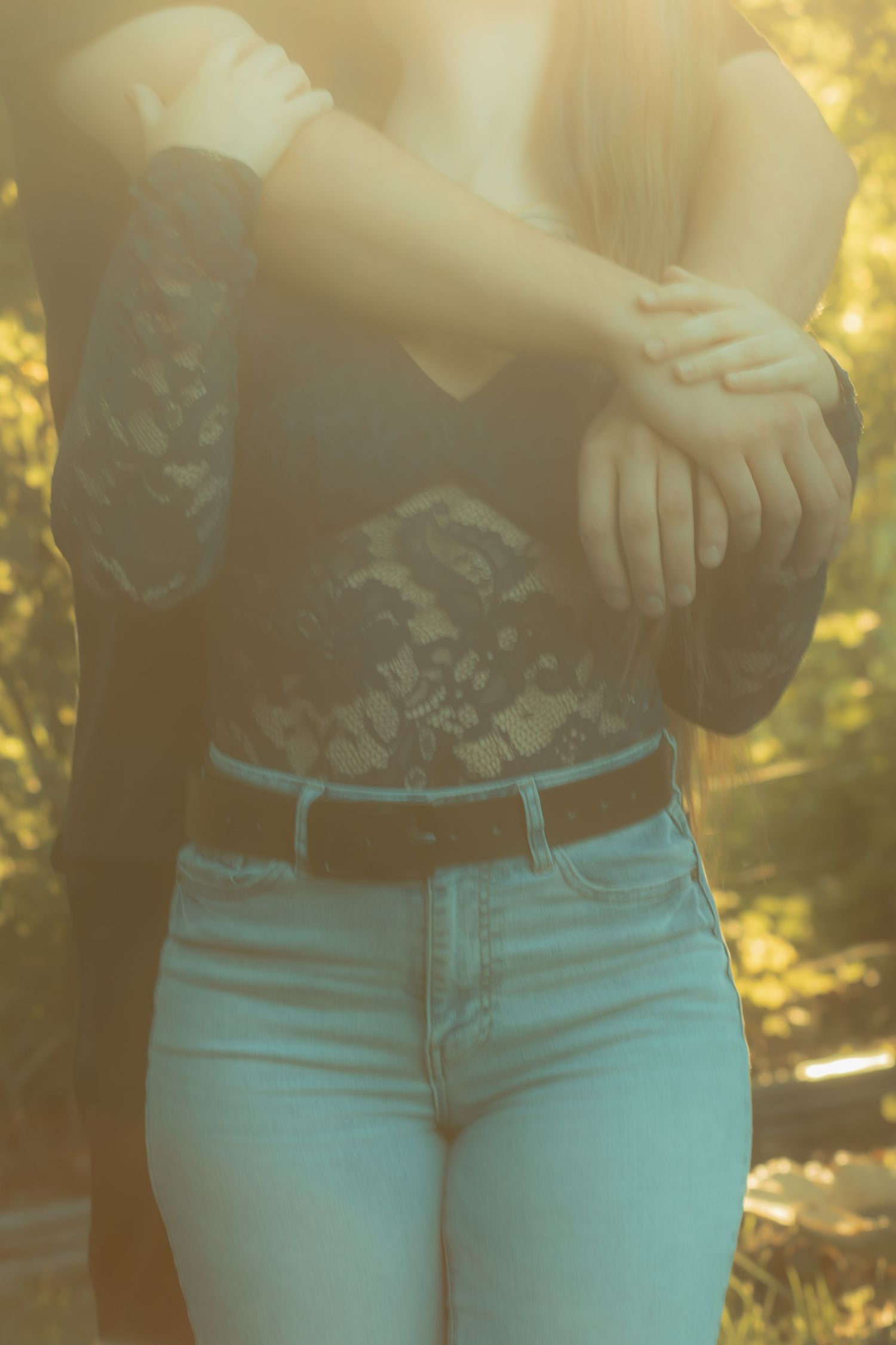 Woman in lace top, black belt, and light blue jeans crossing arms outdoors in warm golden sunlight.