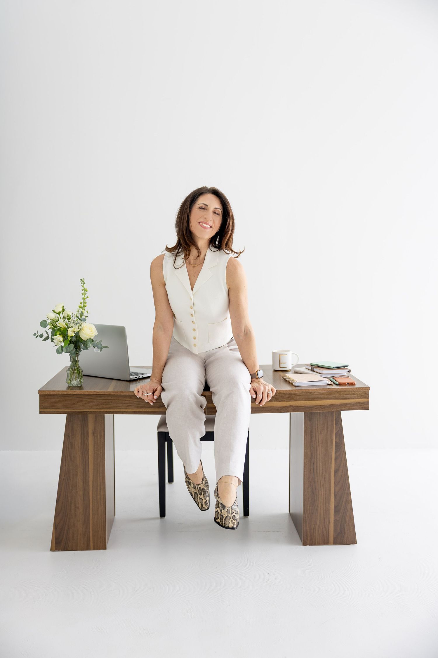 Woman in white sleeveless outfit sitting on a wooden desk in a bright minimalist office studio.