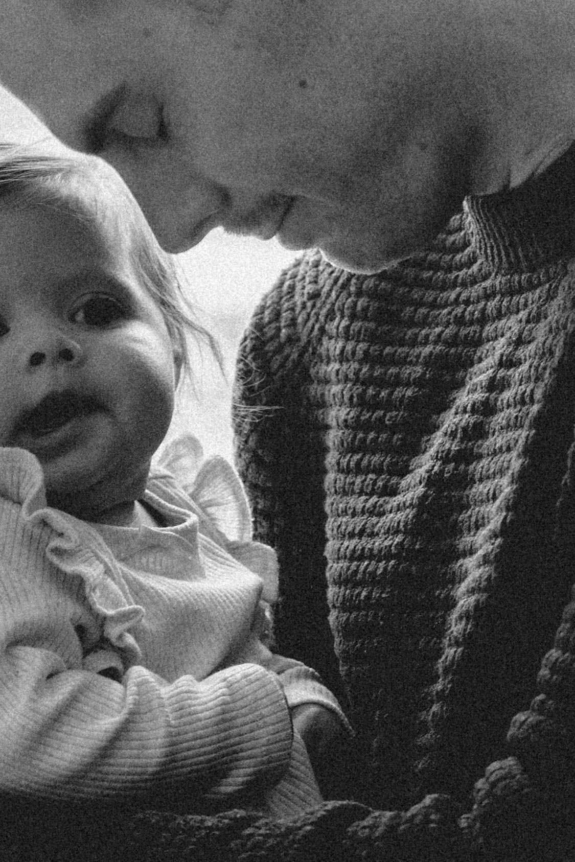 A black and white vintage photograph showing a close-up moment between a baby in ruffled clothing and someone in a knit sweater.