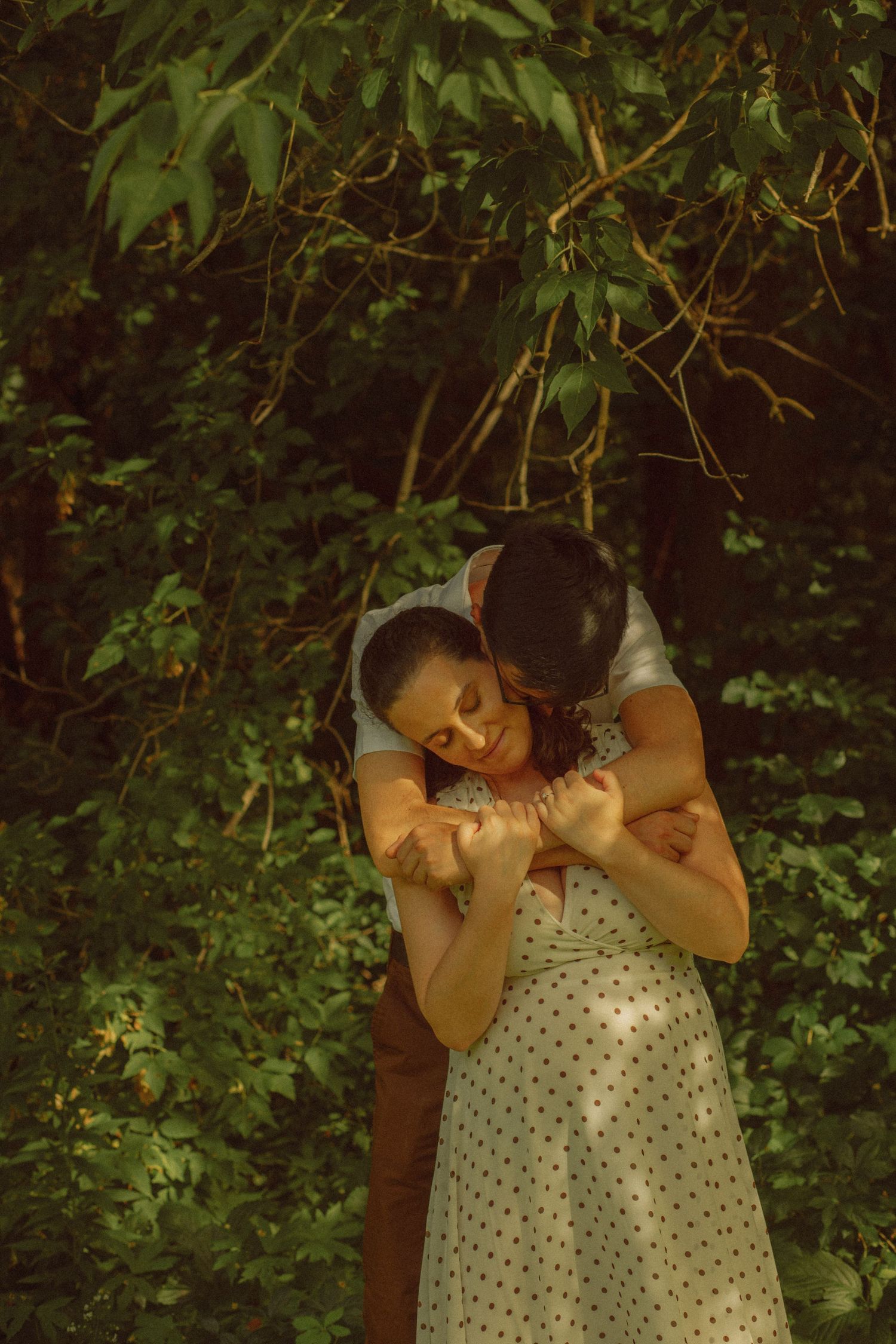 A couple embraces tenderly in a lush green forest, the man holding the woman from behind in soft golden light.