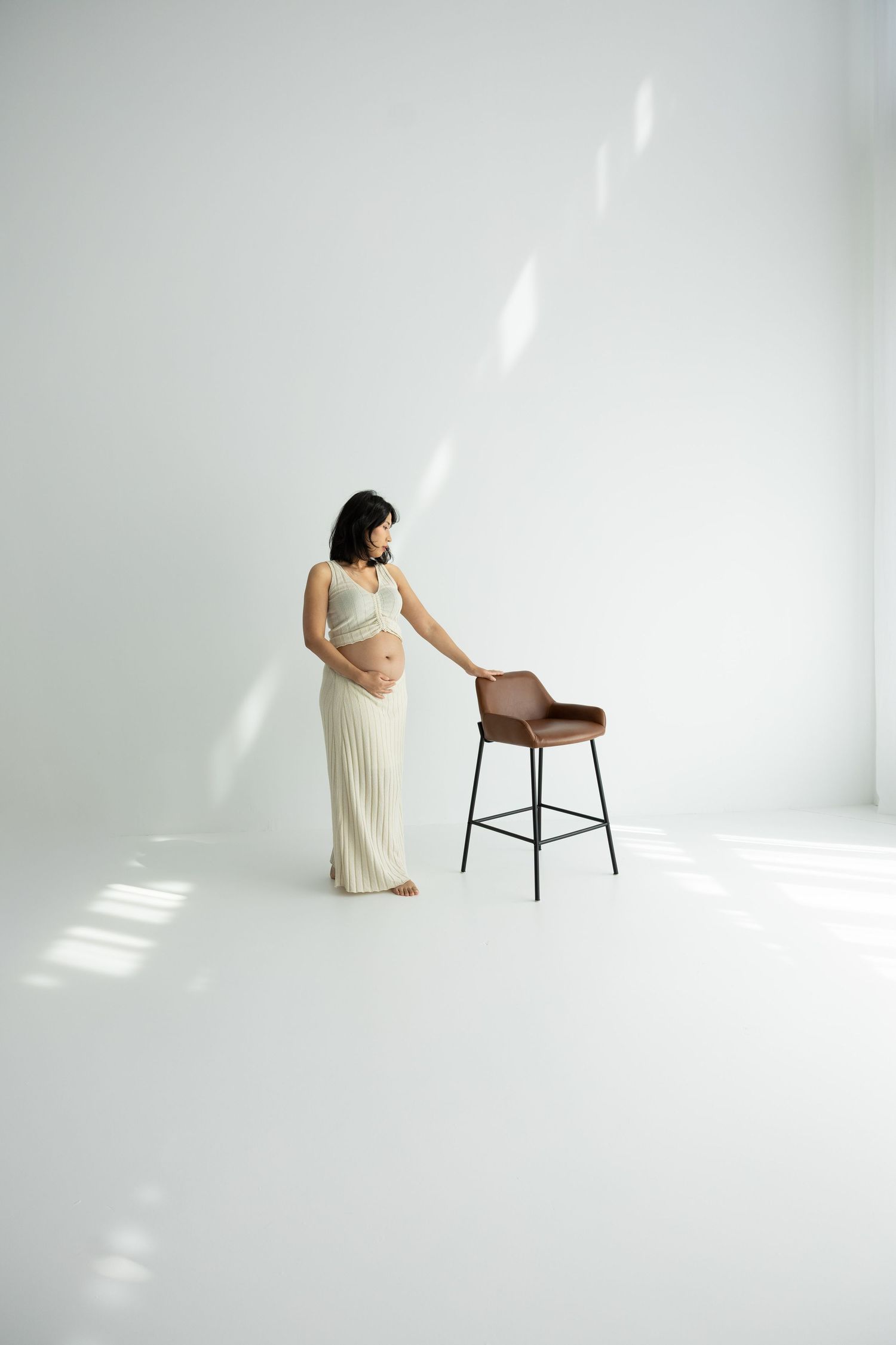 Pregnant woman in cream outfit touching a brown bar stool in a bright white studio with natural light.