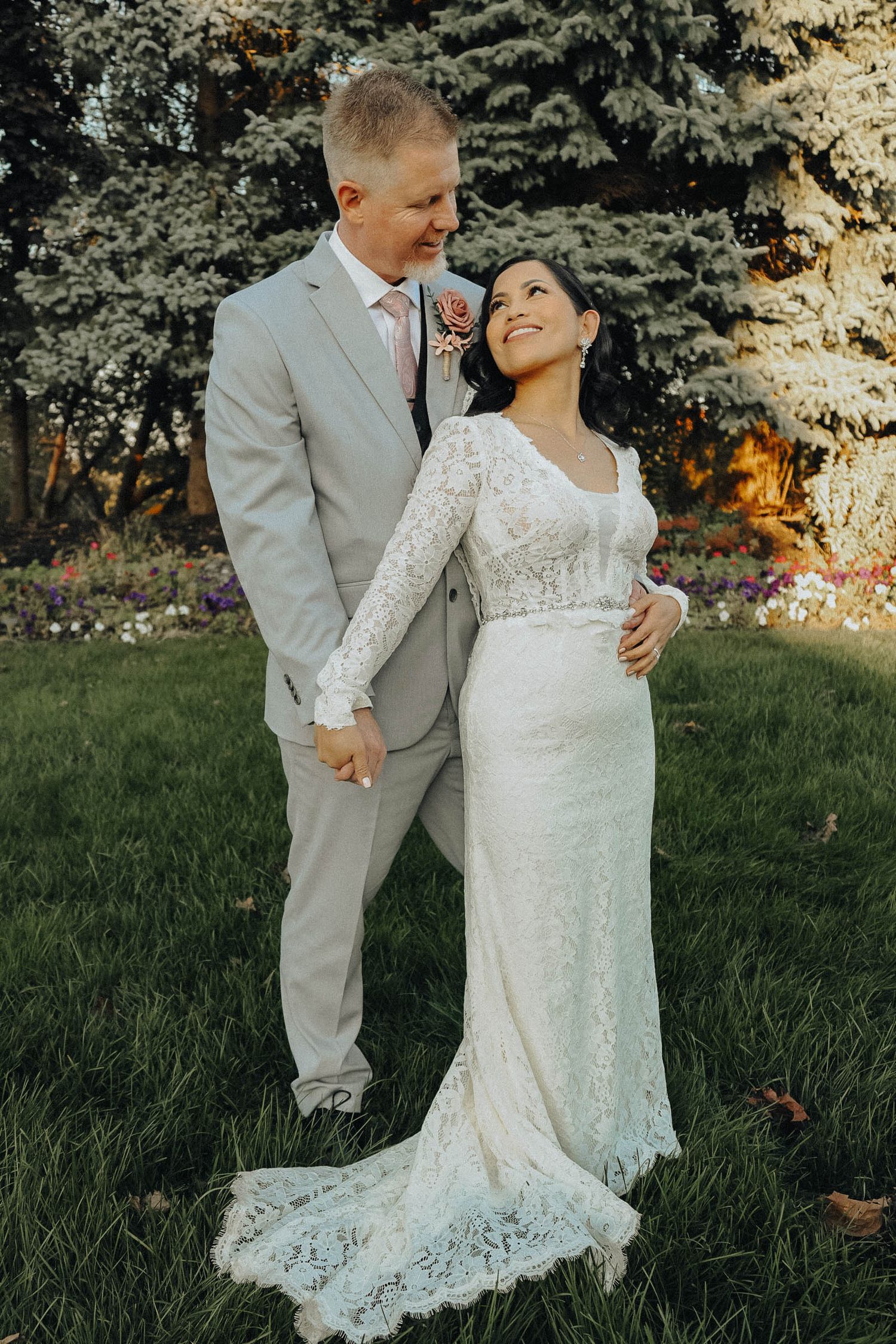 A couple shares an intimate moment in a garden setting wearing a lace wedding dress and grey suit.