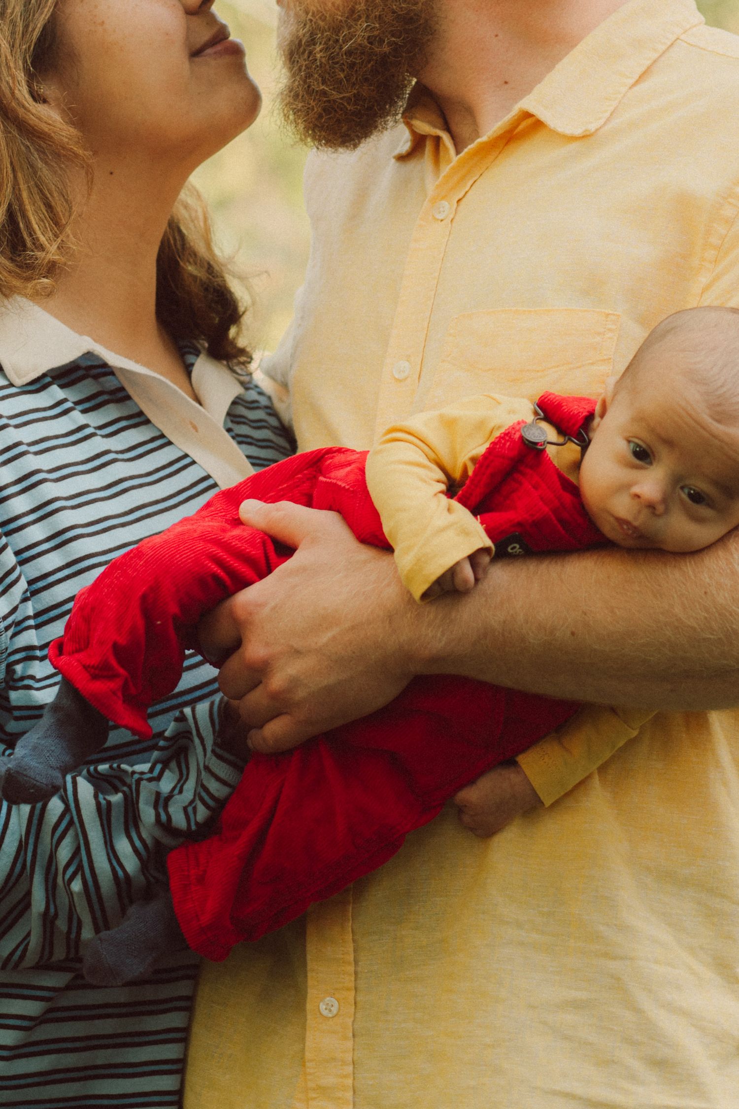 Parents in striped and yellow shirts tenderly hold their newborn baby dressed in a red outfit outdoors.