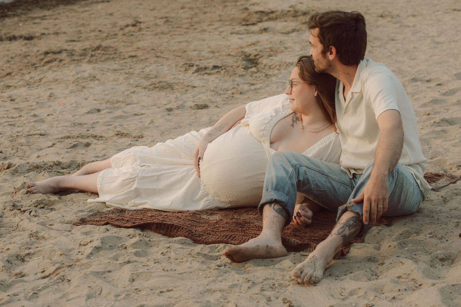 Pregnant couple sitting together on sandy beach, woman in white dress leaning against man in jeans, romantic maternity photo.