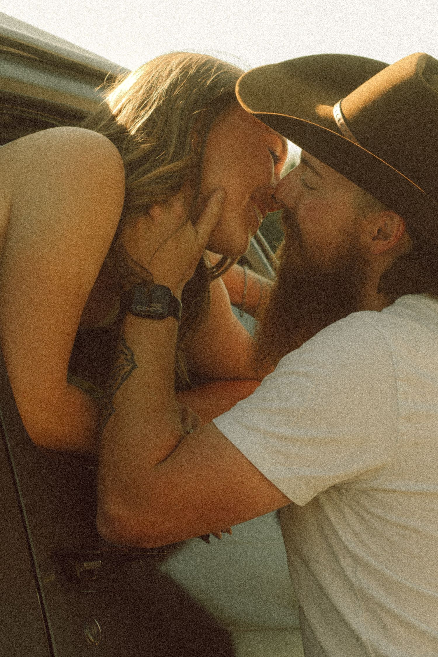A couple shares a romantic kiss near a car window, man wearing a black cowboy hat and white t-shirt, woman with tattoos.