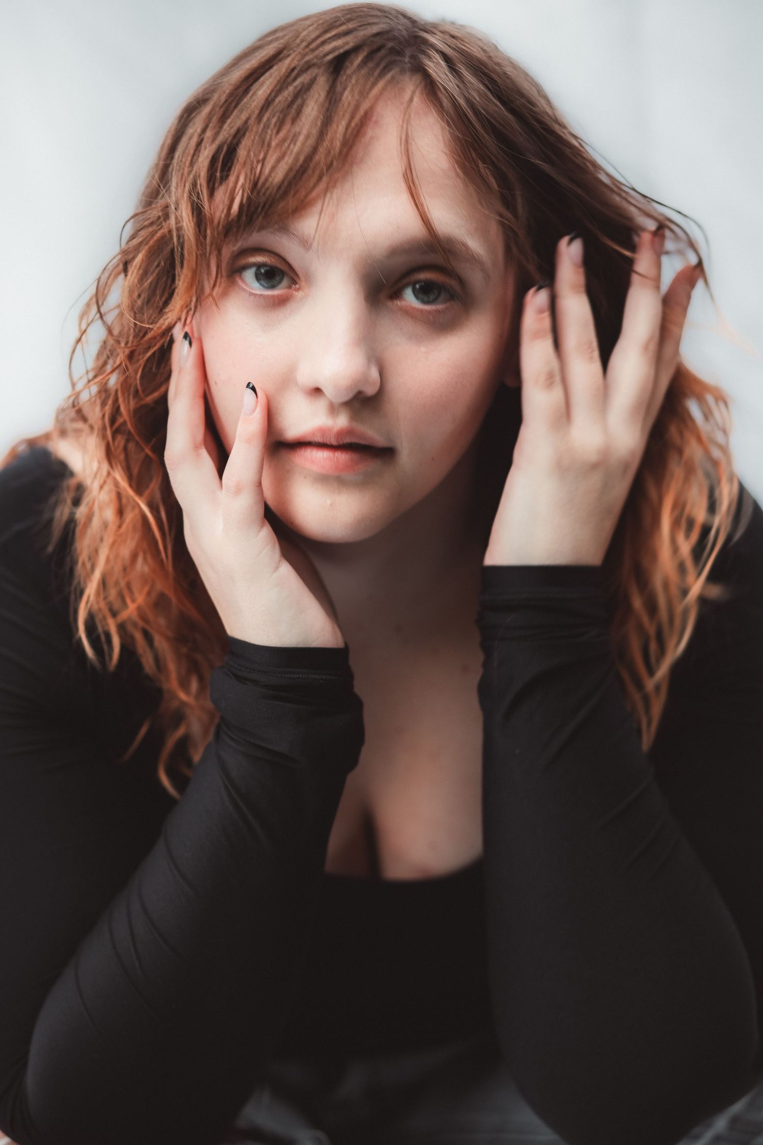 Young woman with red wavy hair in black top, resting face in hands, gazing at camera with a pensive expression.