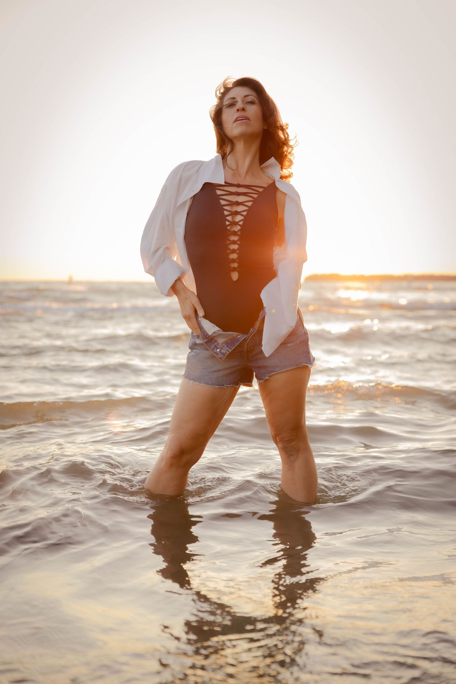 Person wearing black swimsuit and white shirt poses in shallow ocean waters during golden sunset.
