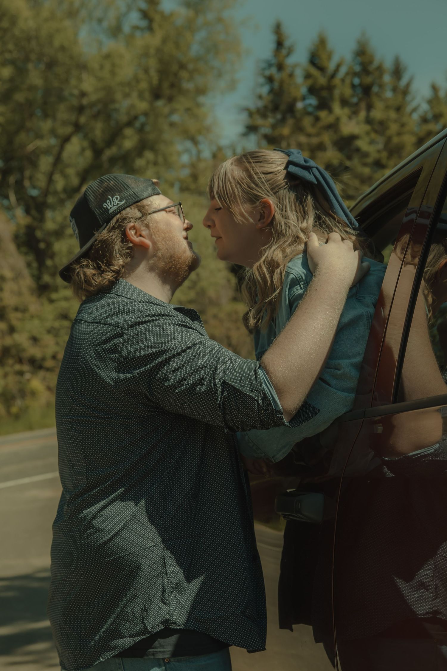 A couple shares a romantic moment leaning toward each other beside a dark vehicle on a tree-lined road.