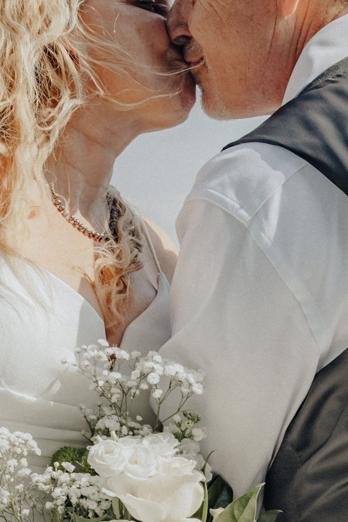 A mature couple shares a romantic kiss at their wedding, with a white rose bouquet in the foreground.