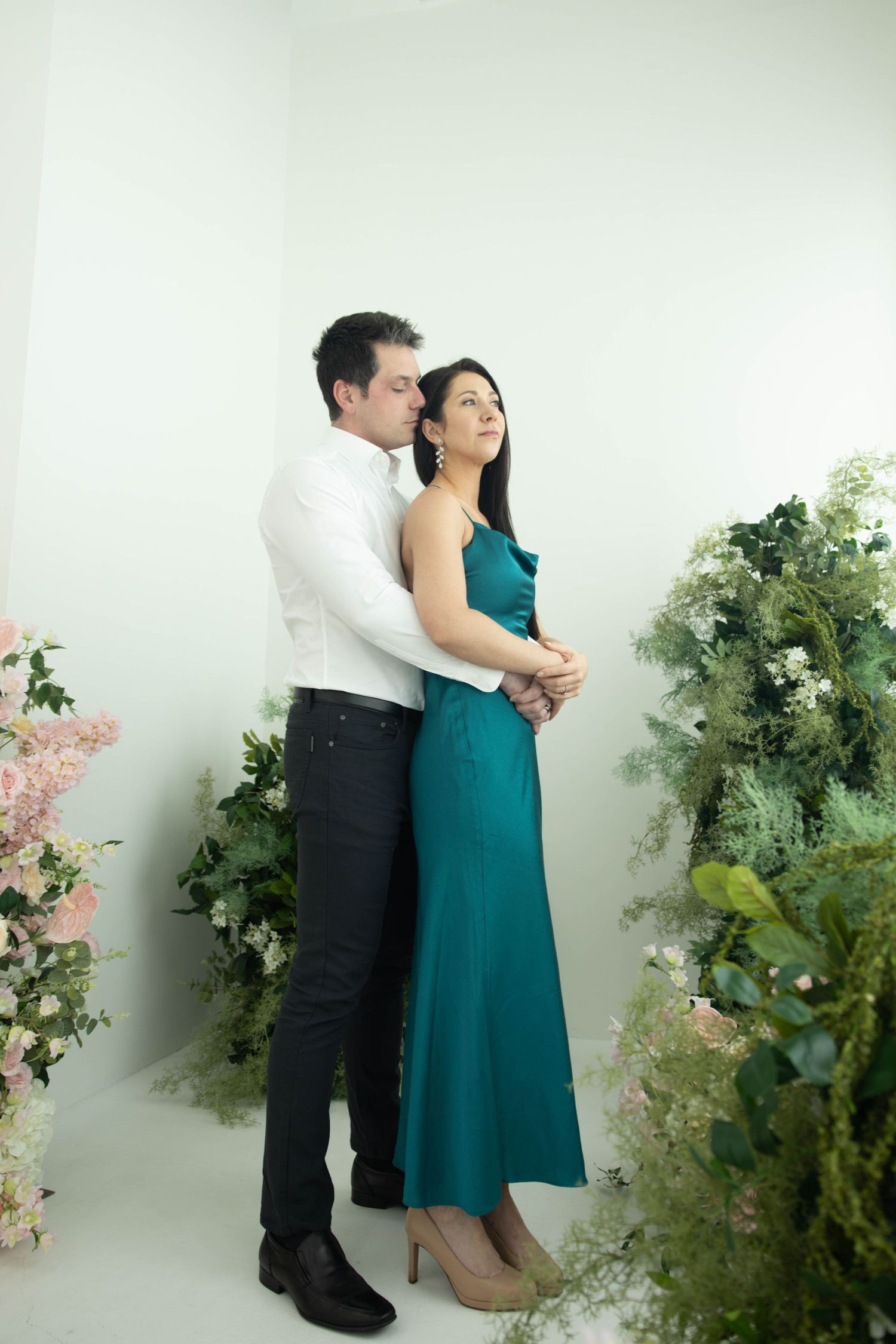 A couple in formal attire shares an intimate moment against a white wall with decorative green foliage and pink flowers.