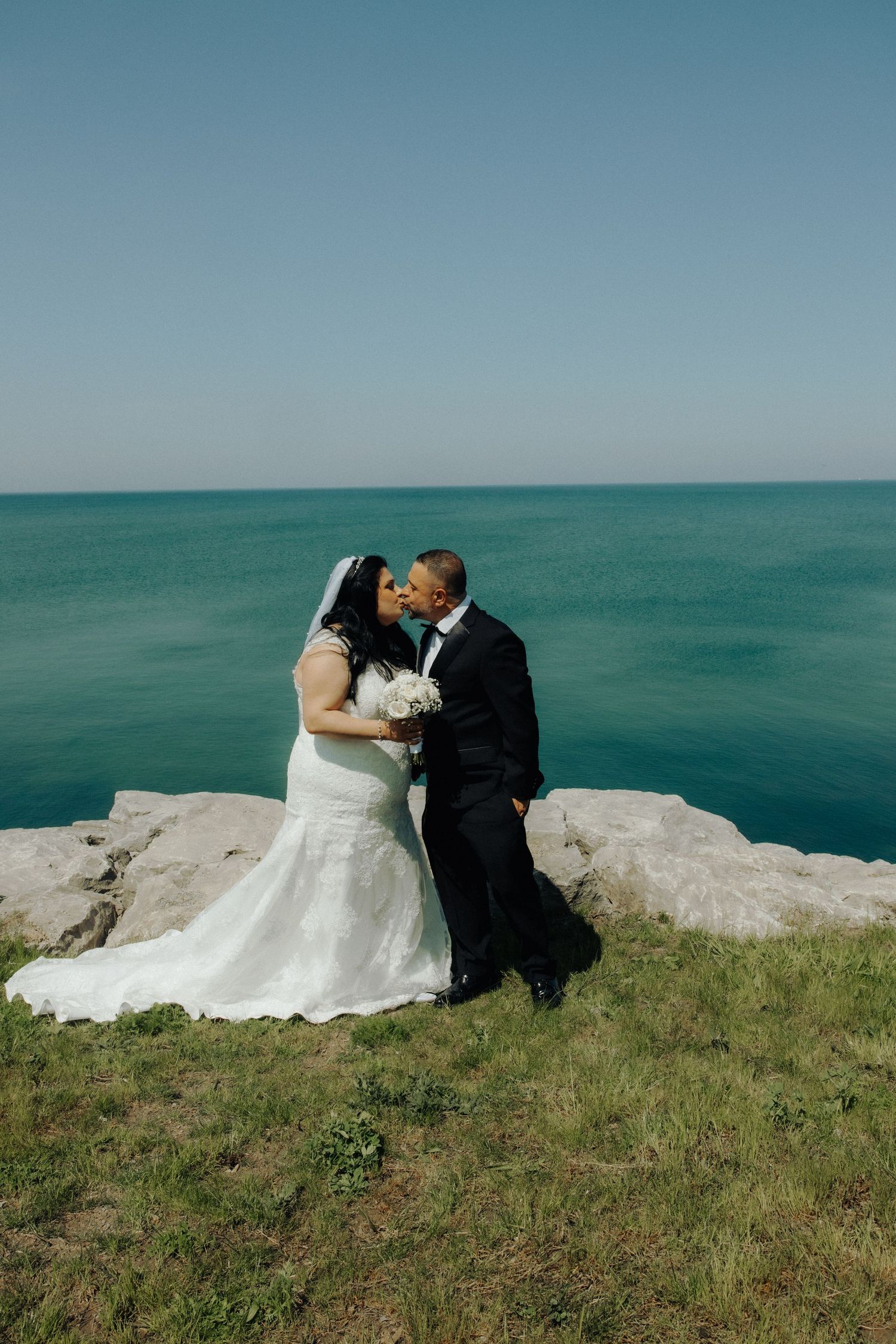 Bride and groom kissing on rocky cliff overlooking turquoise ocean, bride in white lace gown holding bouquet.