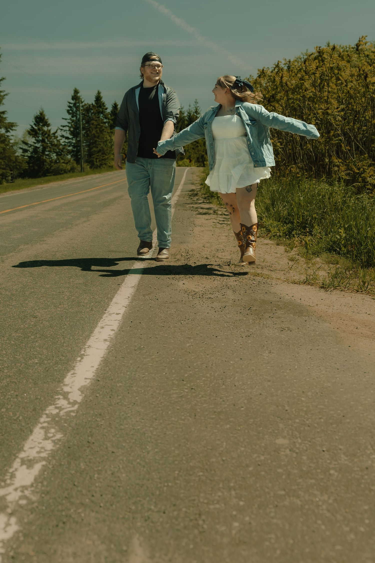 A happy couple walks hand in hand along a scenic rural road surrounded by trees on a sunny day.