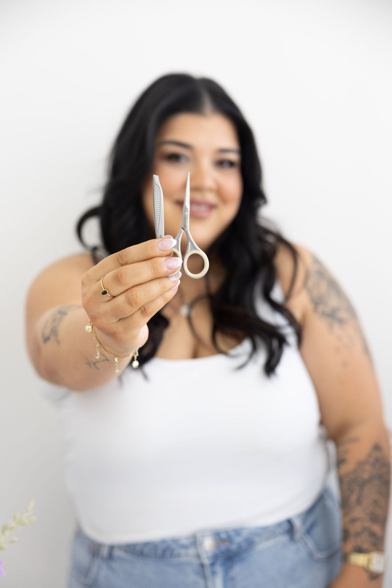 A smiling plus-size woman with tattoos holds hair scissors toward the camera in a bright white studio.