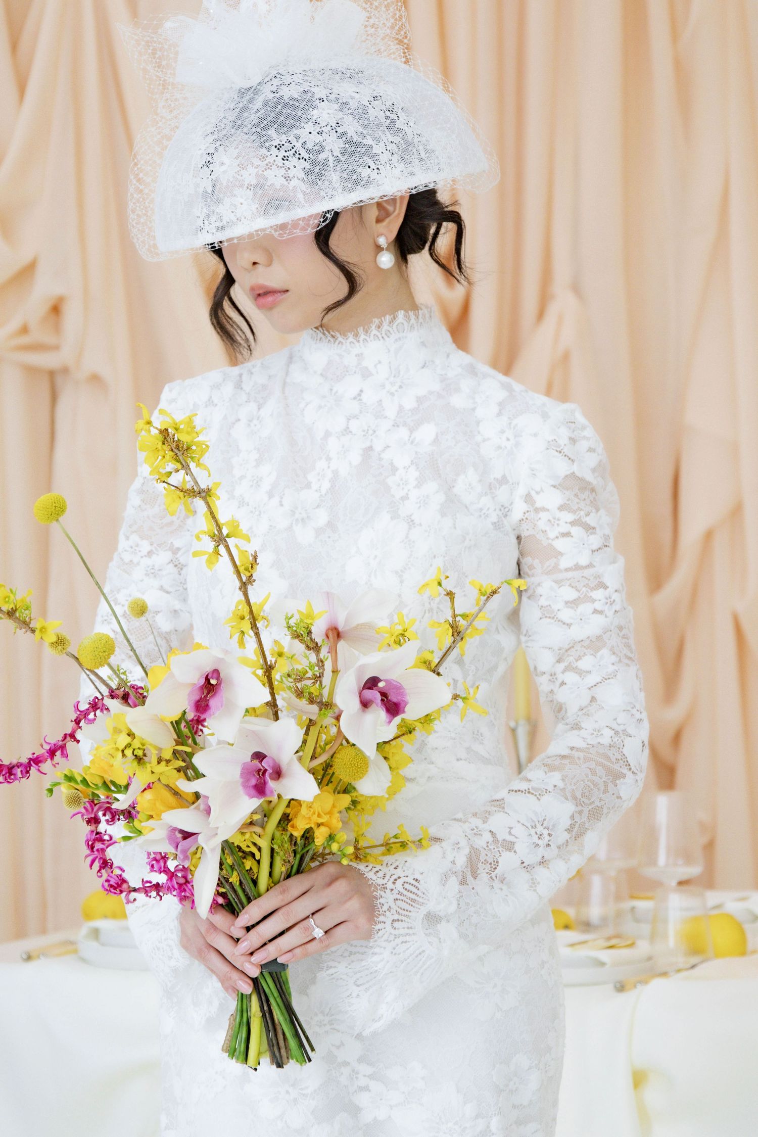 Bride in elegant white lace gown and veil holds a colorful spring bouquet with yellow and pink flowers.