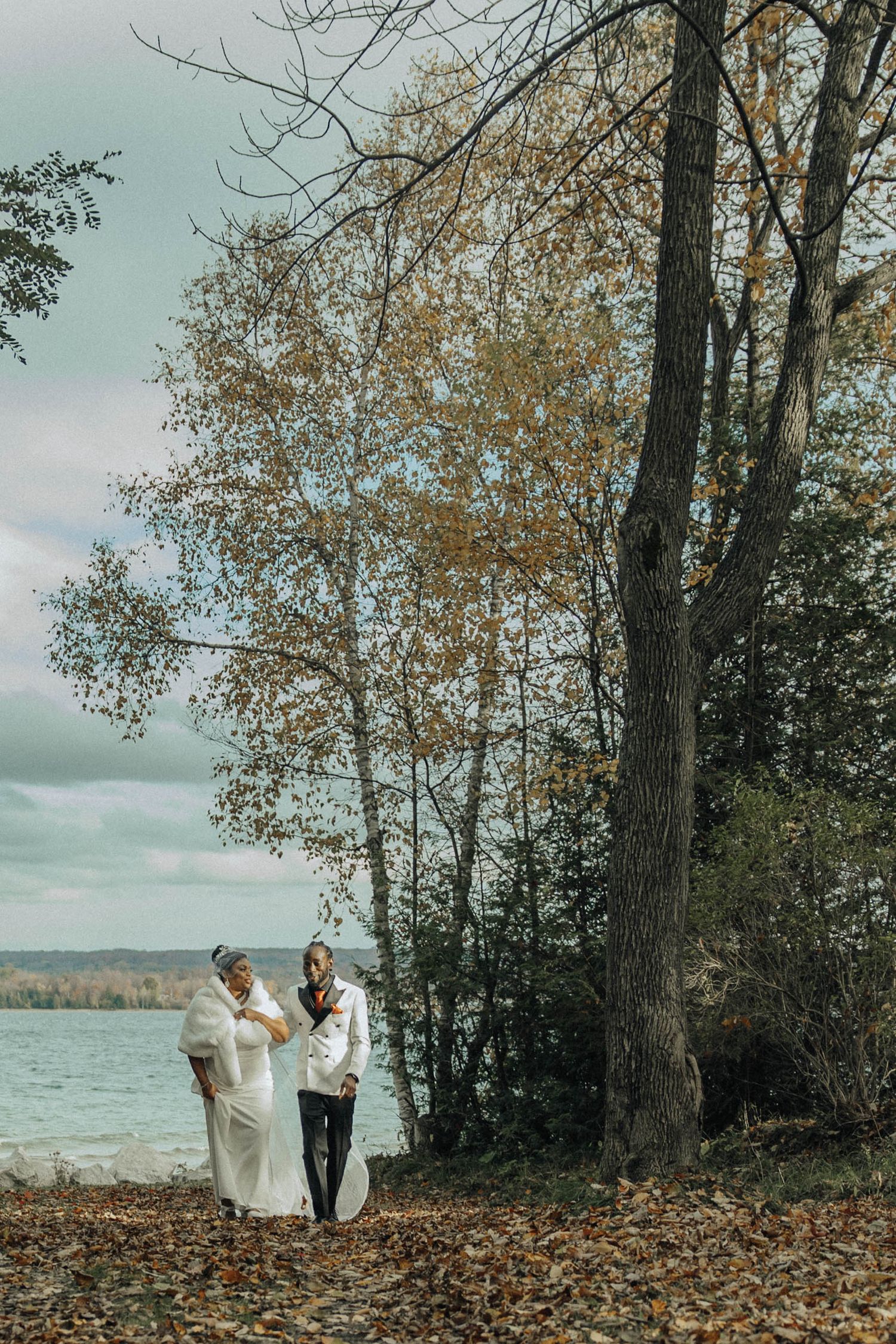 A romantic autumn wedding scene by a lake with tall trees and fallen leaves on the ground.