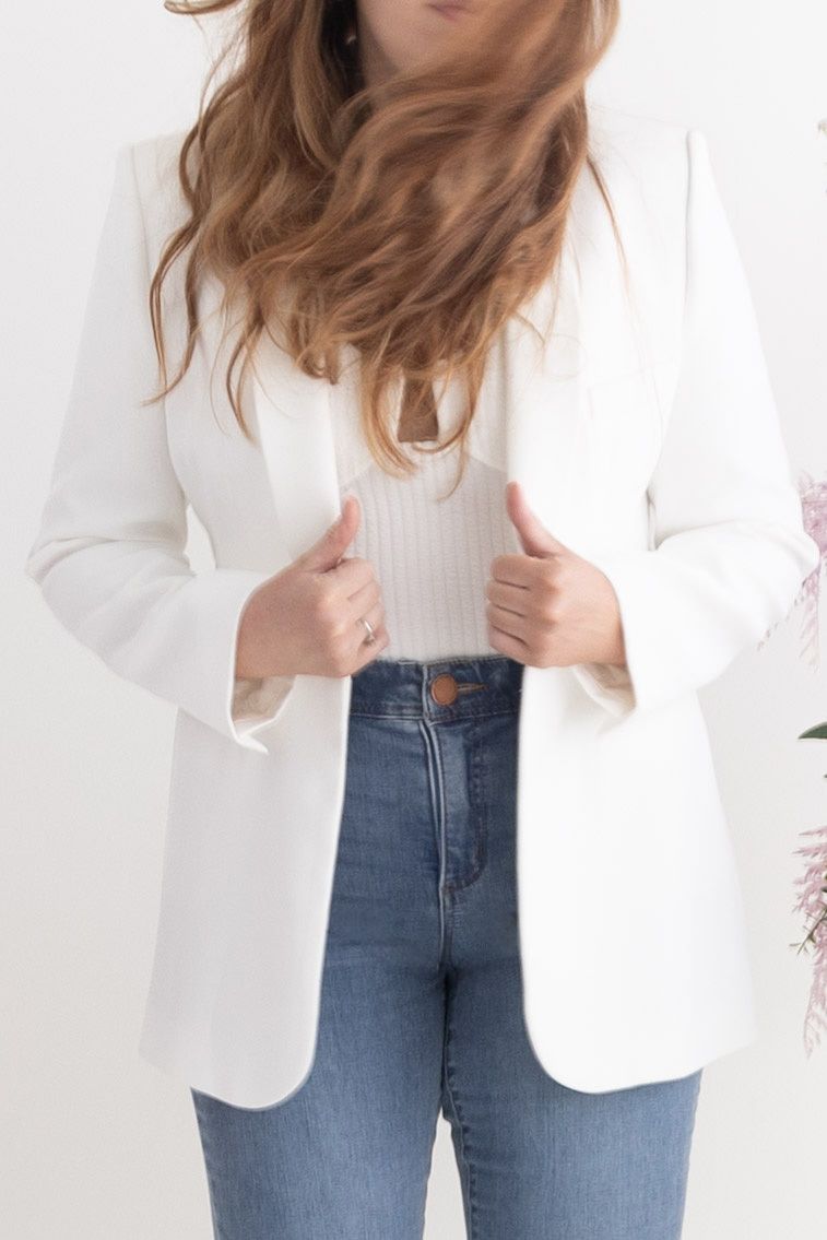 Woman wearing a white blazer over a ribbed top with high-waisted blue jeans, posing against a white background.