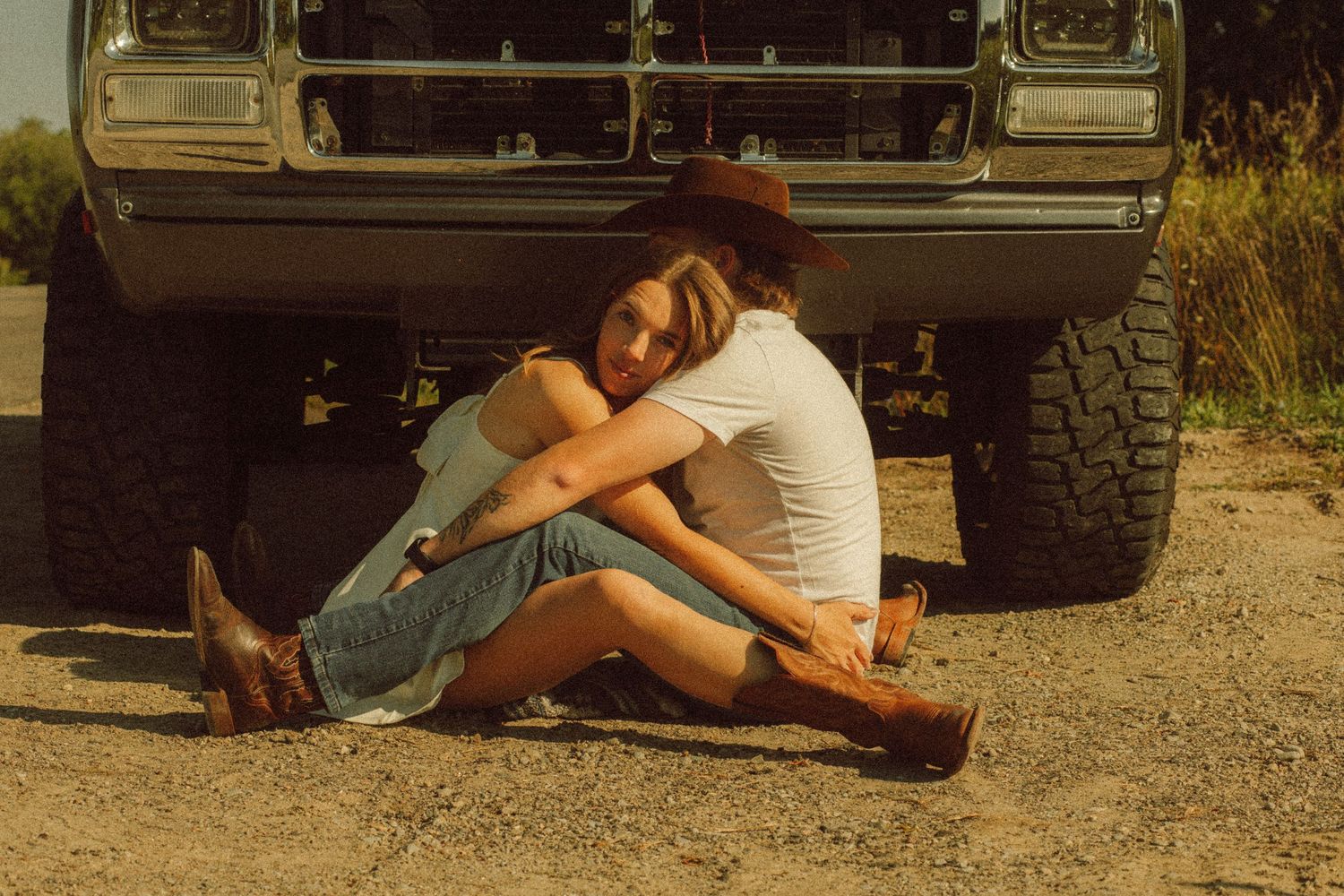 Young woman in cowboy hat and denim skirt sitting against a vintage truck on a dirt road.