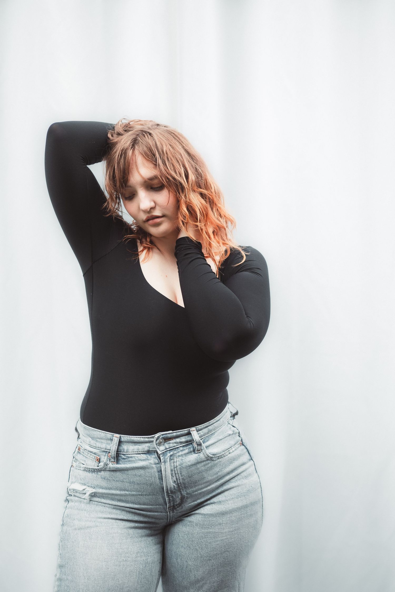 Curly red-haired woman in black V-neck bodysuit and light wash jeans posing against a white curtain backdrop.