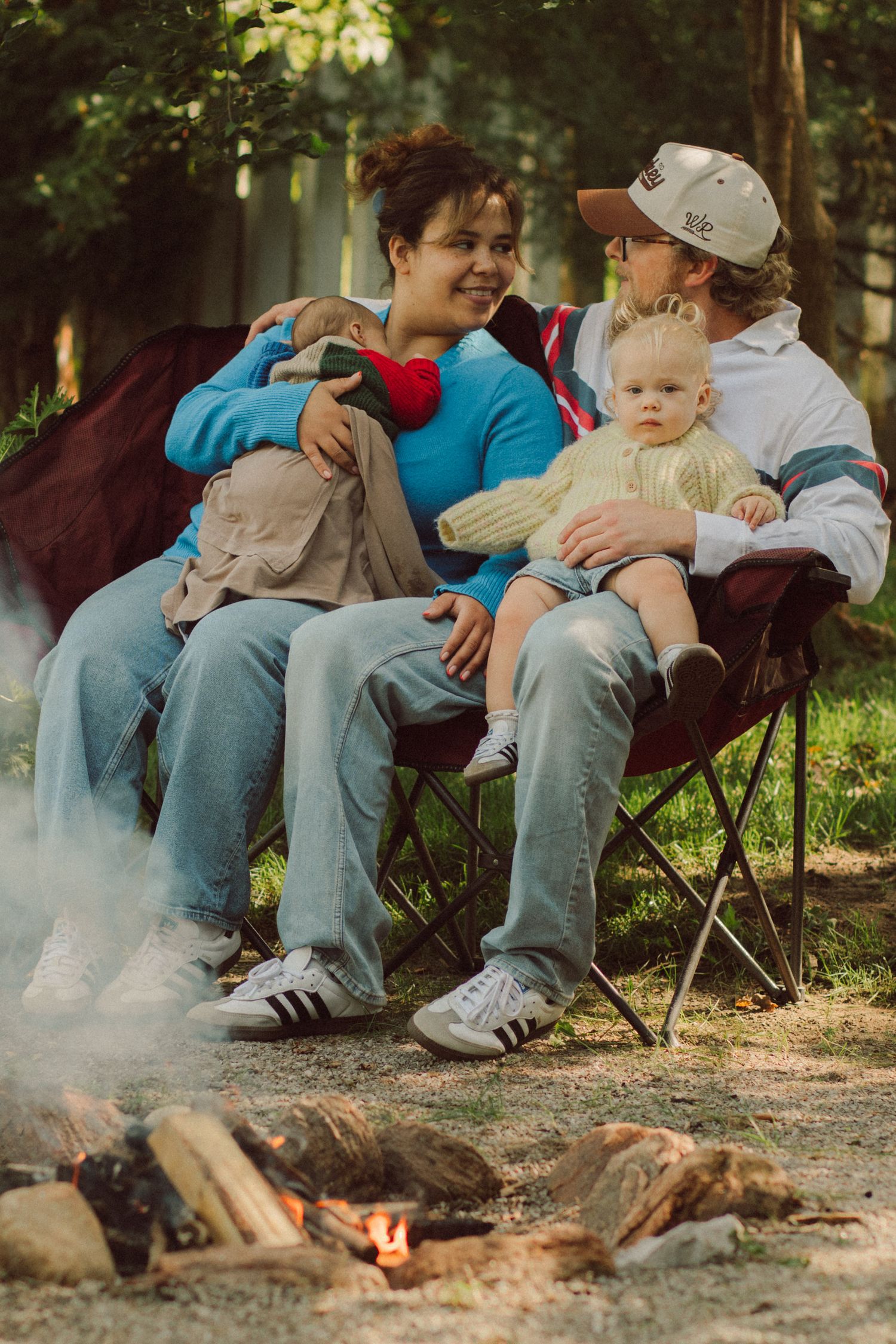 Family sitting around a campfire outdoors, with two adults holding babies in folding chairs among trees.