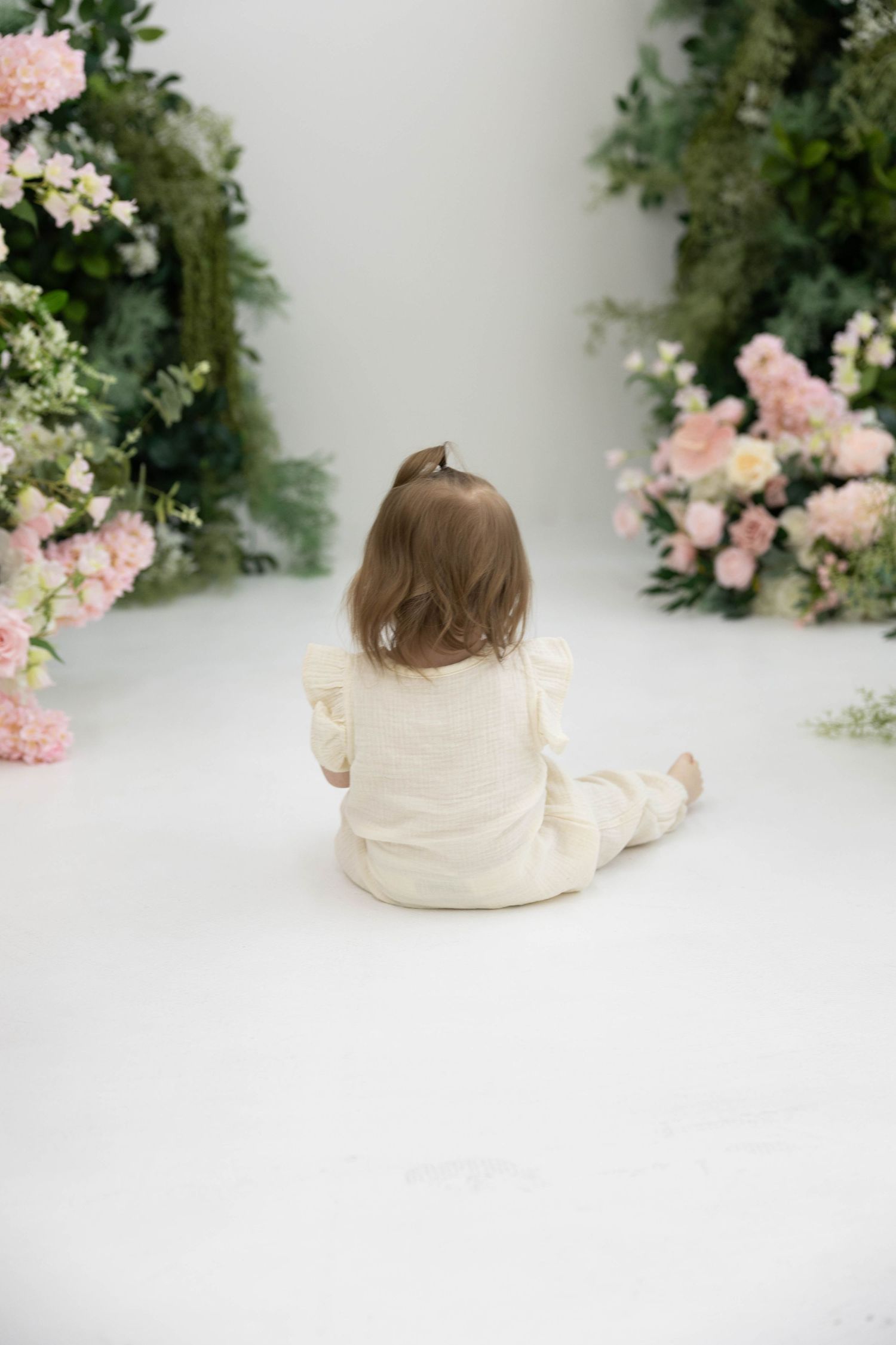 A small child in cream colored clothing sits on white floor surrounded by pink floral arrangements and greenery.
