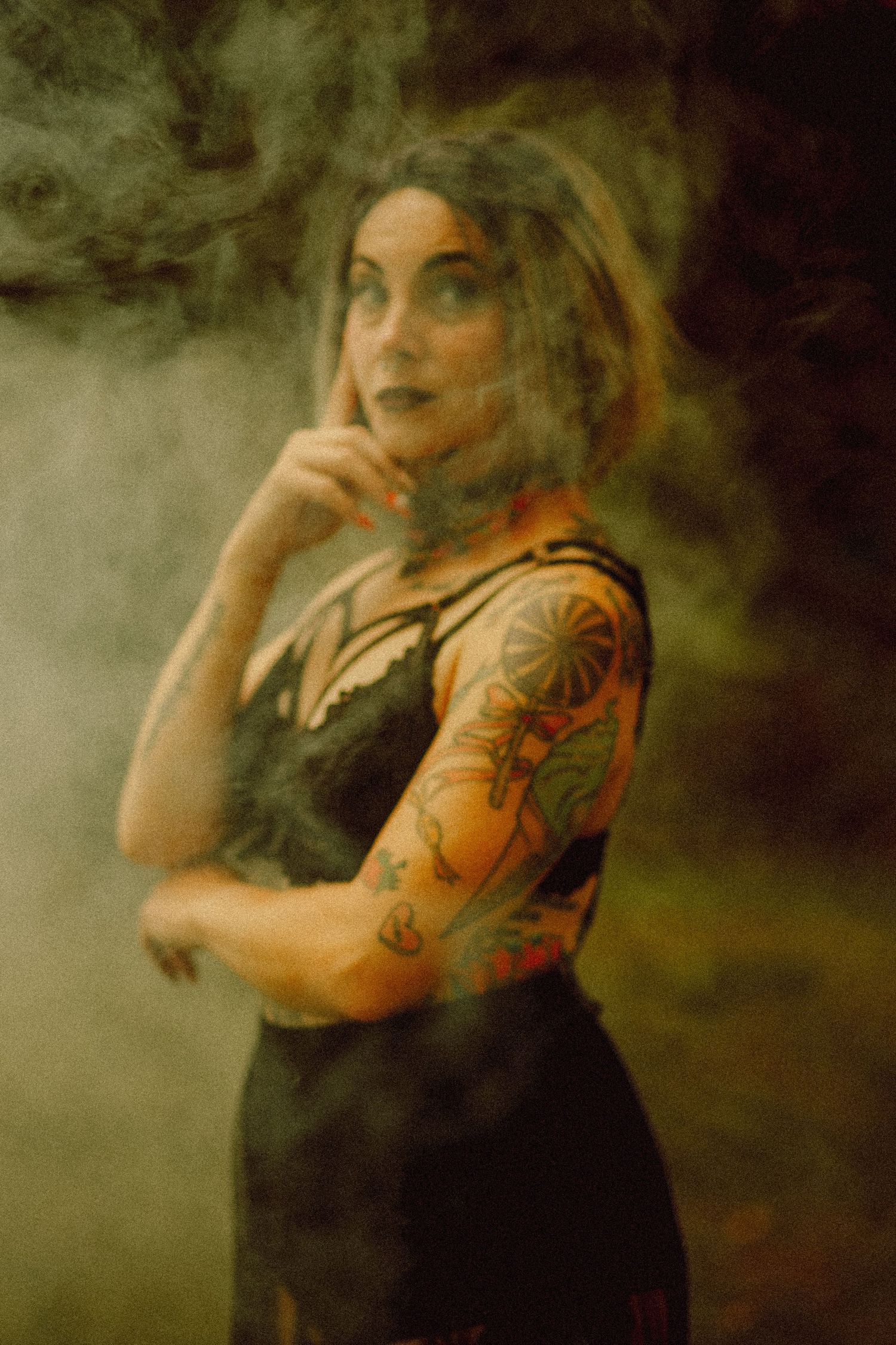 Tattooed woman in black dress poses dramatically in smoky atmosphere, showcasing colorful sleeve tattoos.