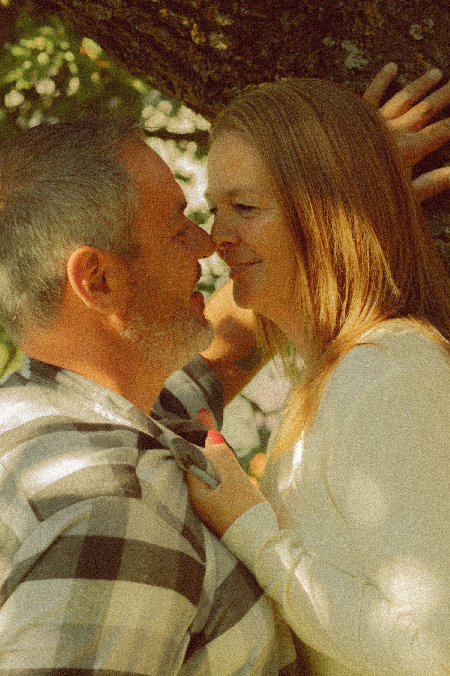 A couple shares a romantic moment nose-to-nose under a tree, the man in plaid and woman in white top smiling softly.