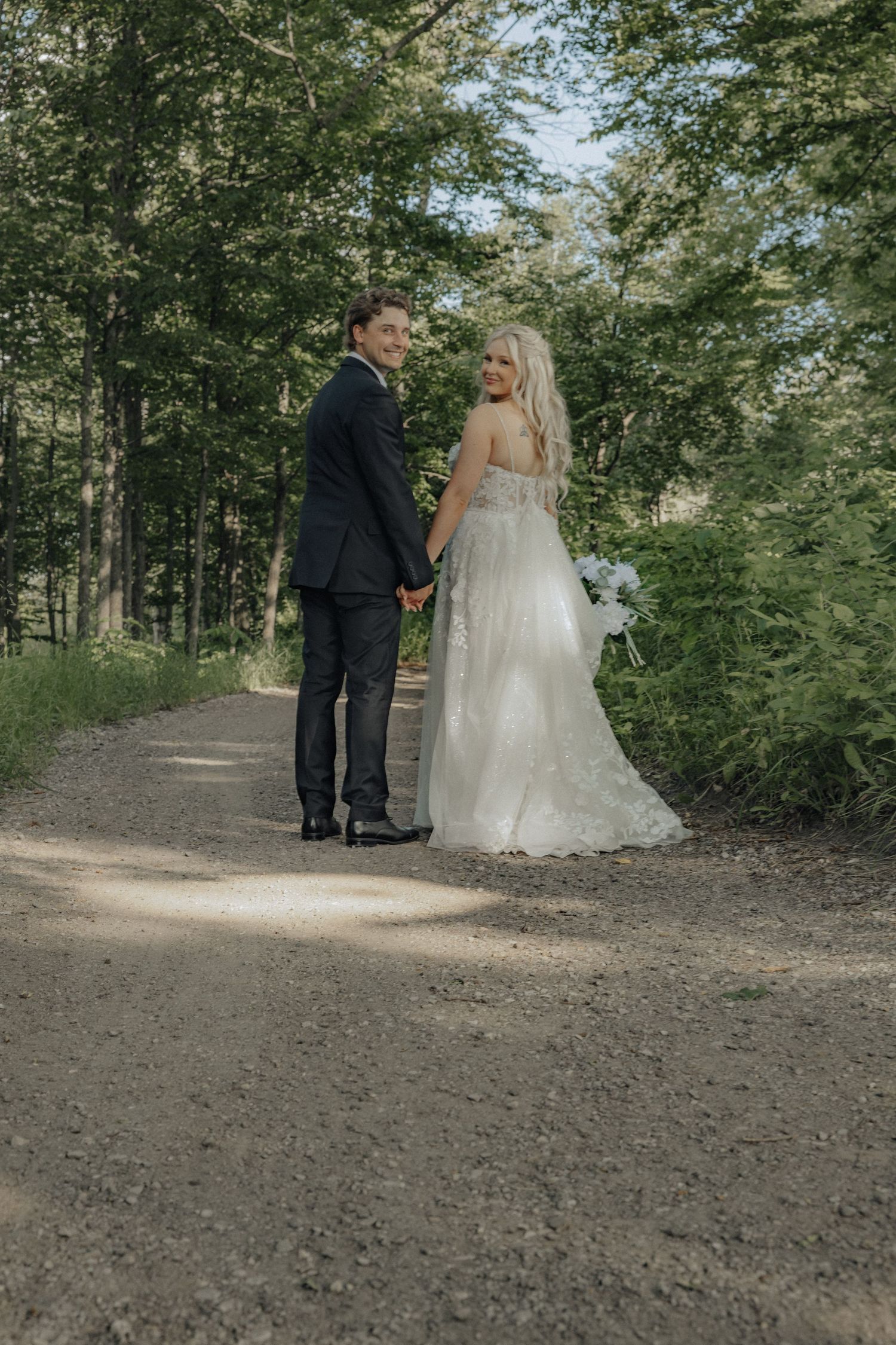 Bride and groom holding hands on a forest path, smiling back at camera, bride in white gown holding blue bouquet.