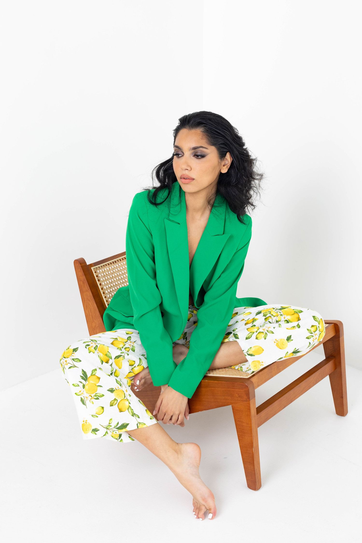 Model poses on wooden chair wearing vibrant green blazer and floral pants against white backdrop.