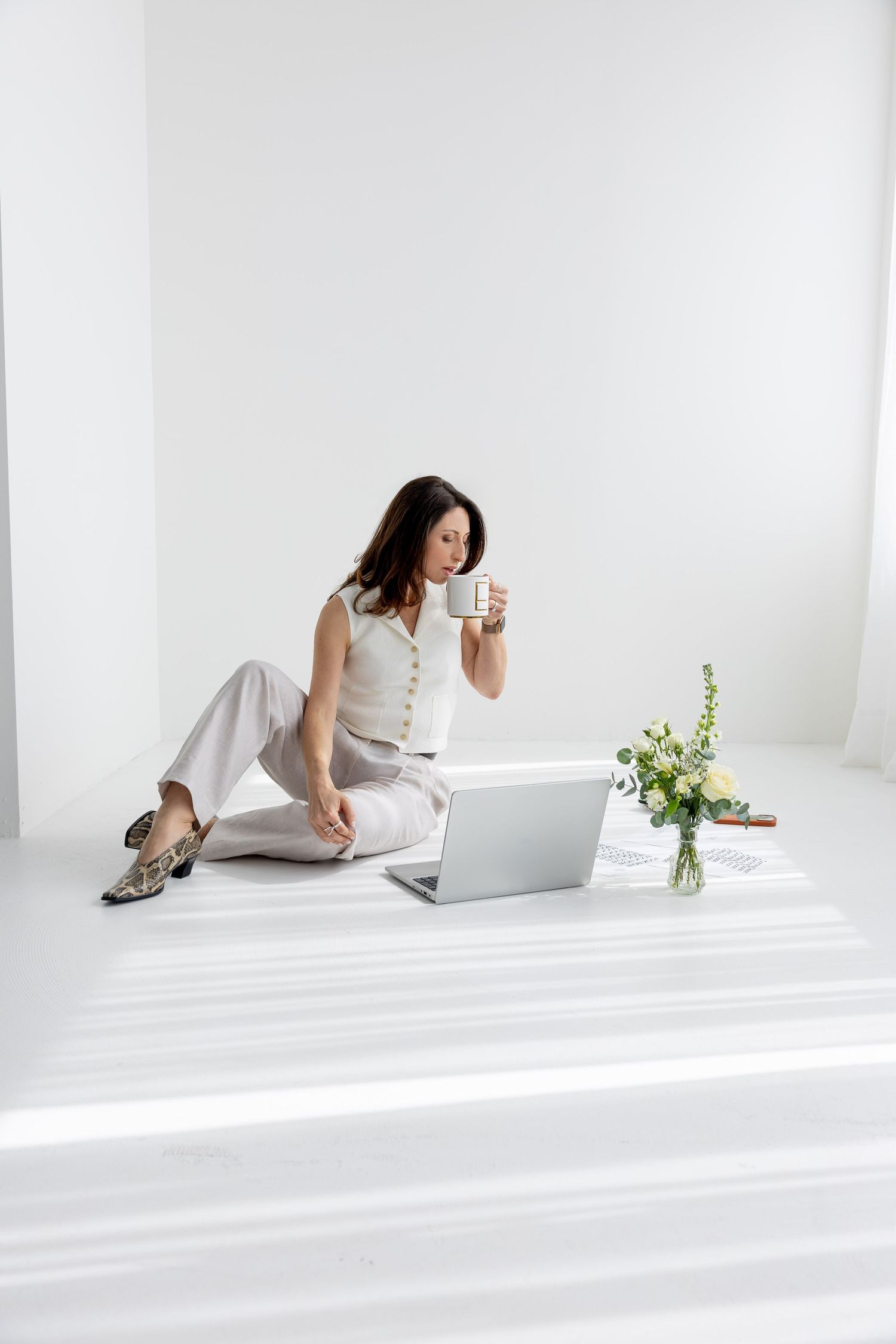 Woman in white outfit sitting on floor with laptop, sipping coffee near fresh flowers in bright minimalist space.