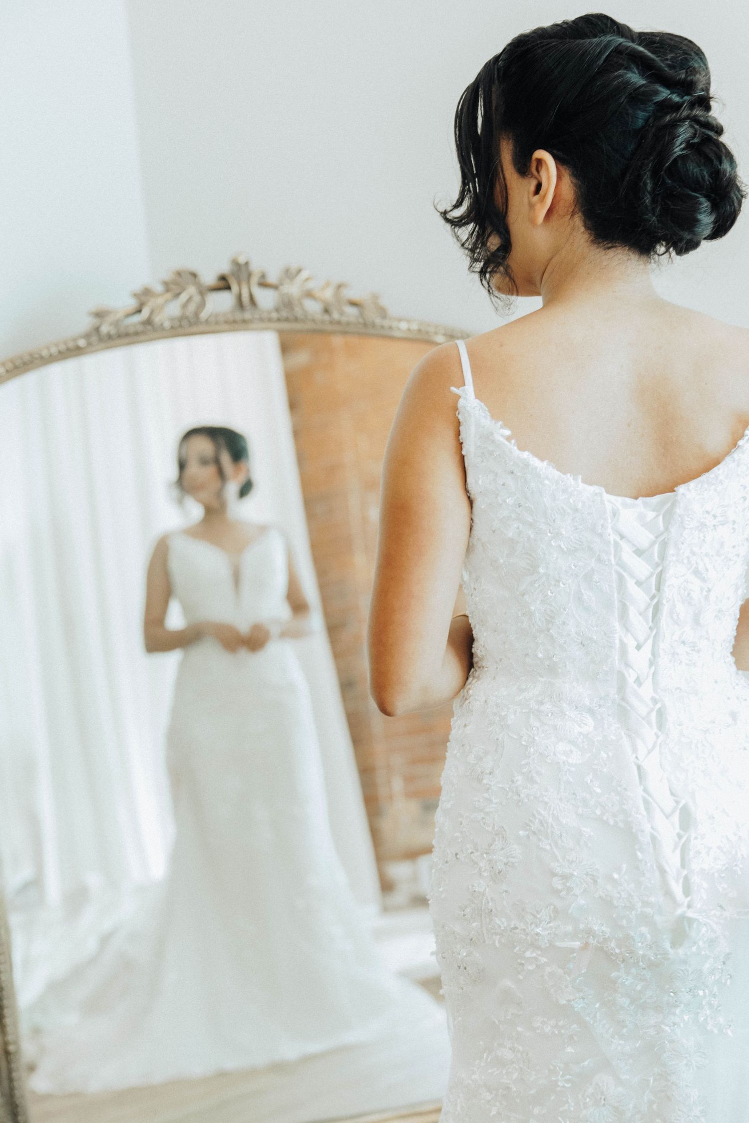 A bride in a white lace wedding dress looks at her reflection in an ornate vintage mirror.