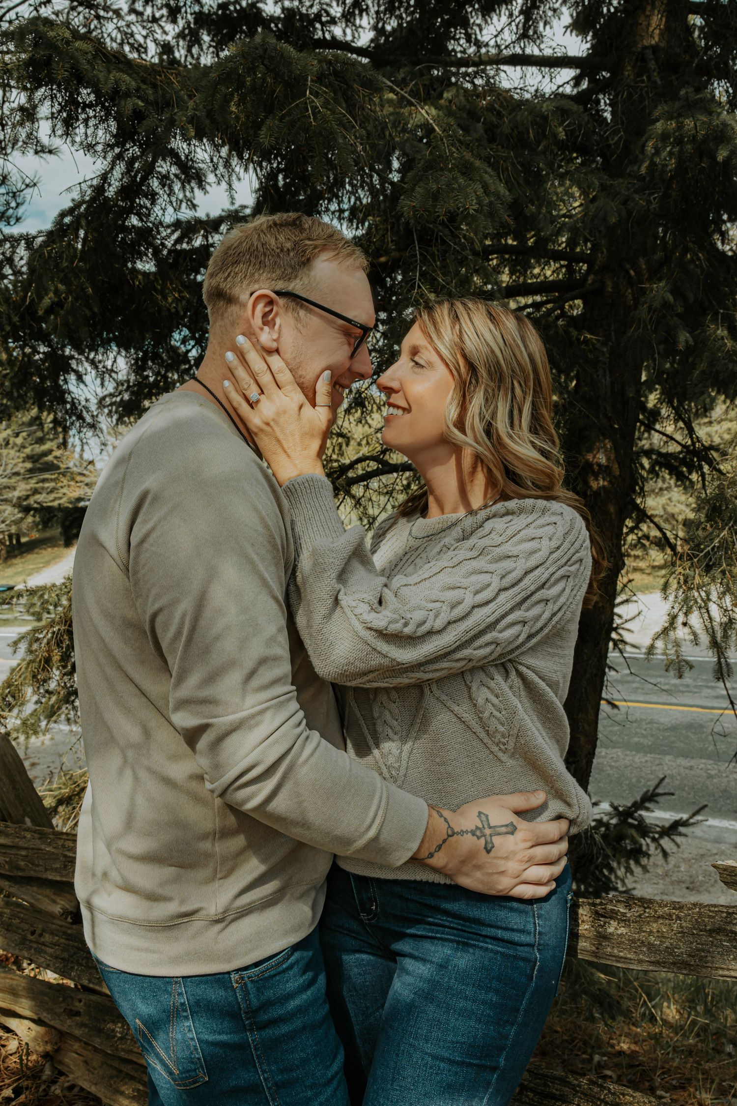 Couple sharing a romantic moment outdoors near pine trees, wearing matching gray sweaters and jeans.