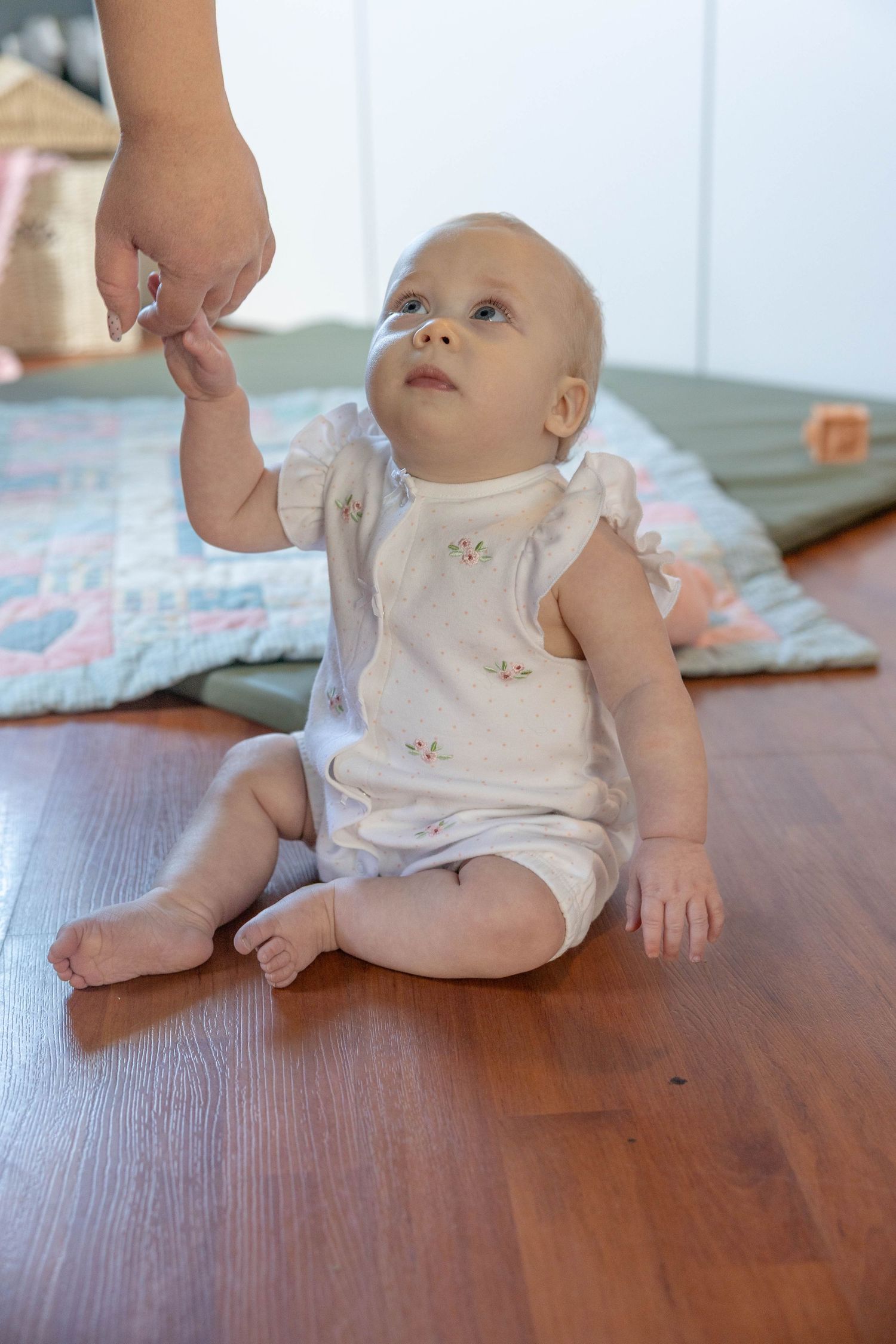 Baby in white romper sits on hardwood floor reaching up toward someone's hand while looking upward.